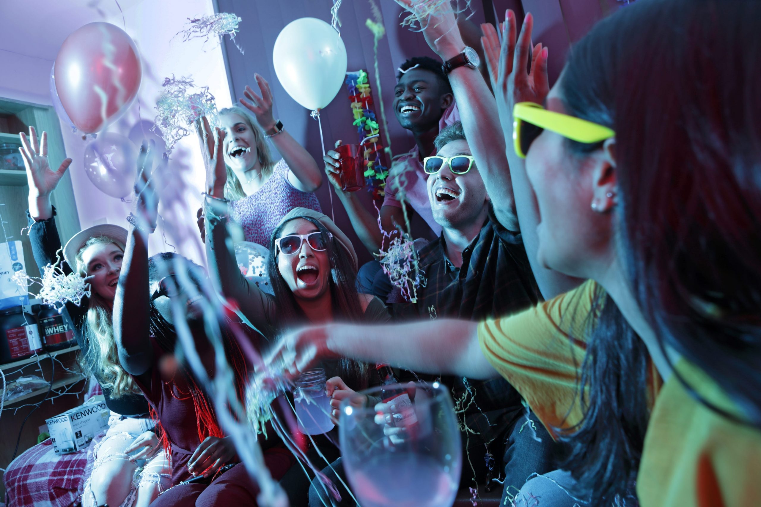 Young people wearing neon glasses dance and throw balloons around at a house party.