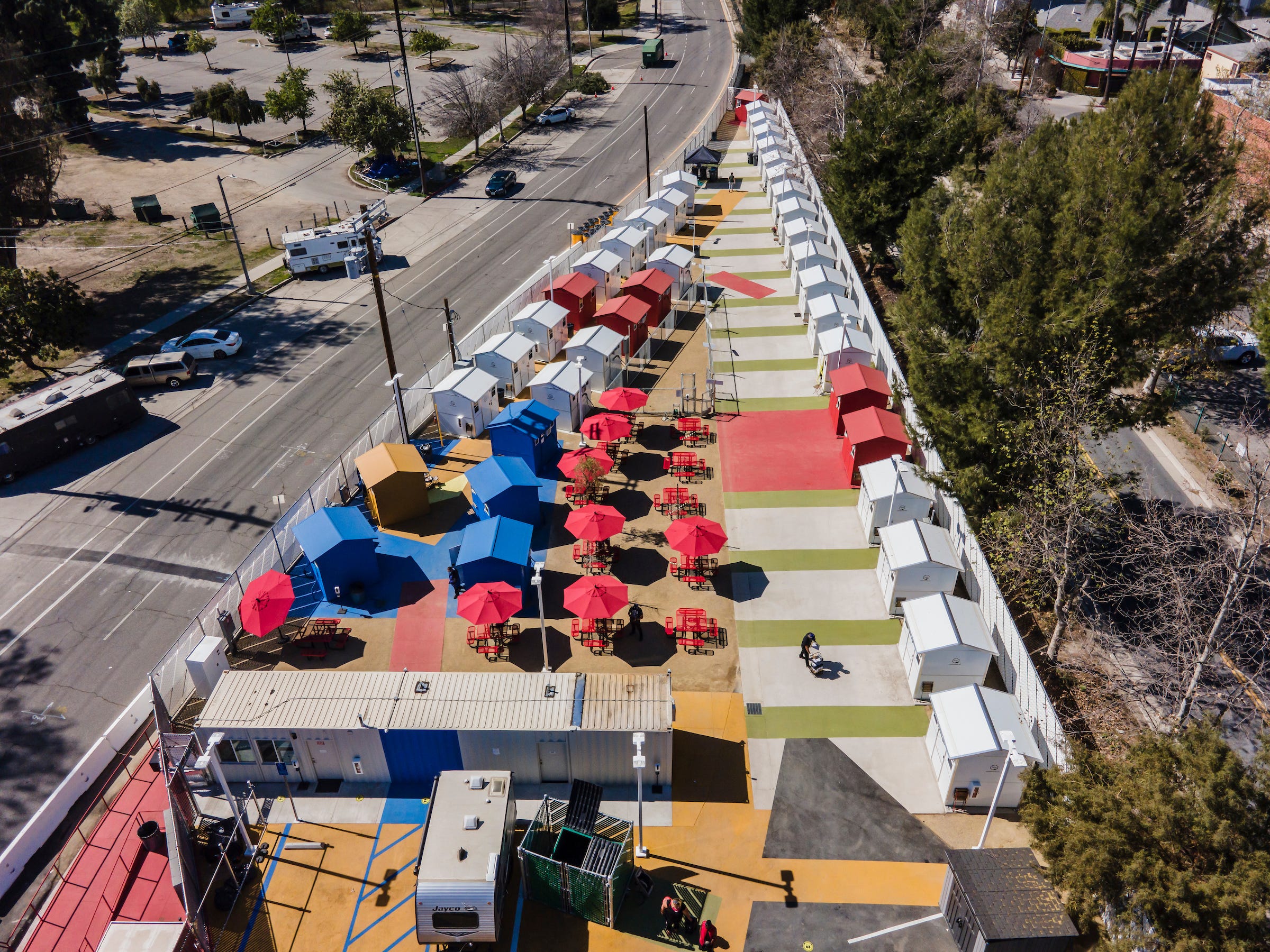 A lot adjacent to a main road is covered with umbrellas and small structures in red, yellow, and blue