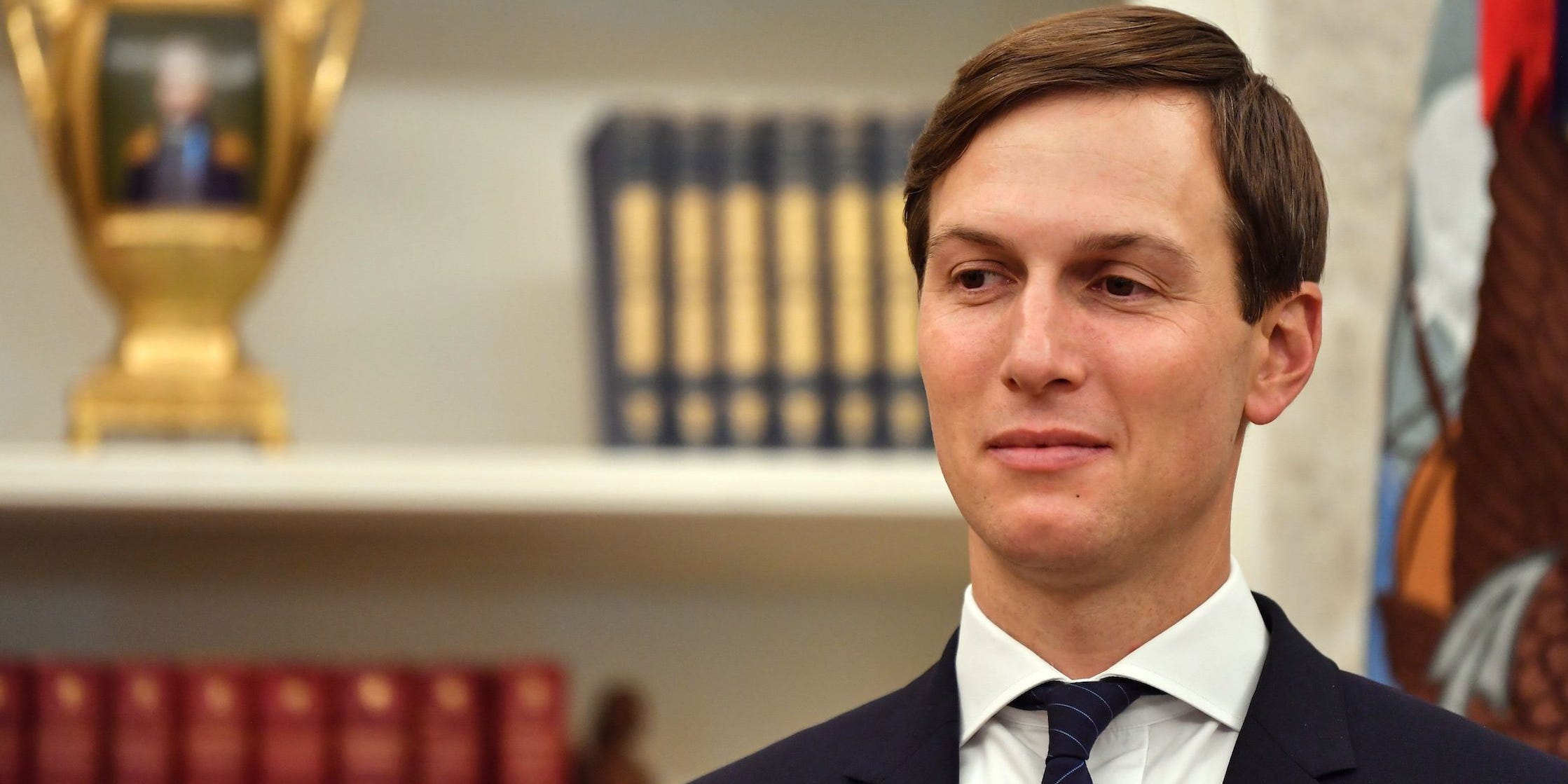 Jared Kushner stands in the oval office in front of a bookshelf with a golden chalice and two collections.