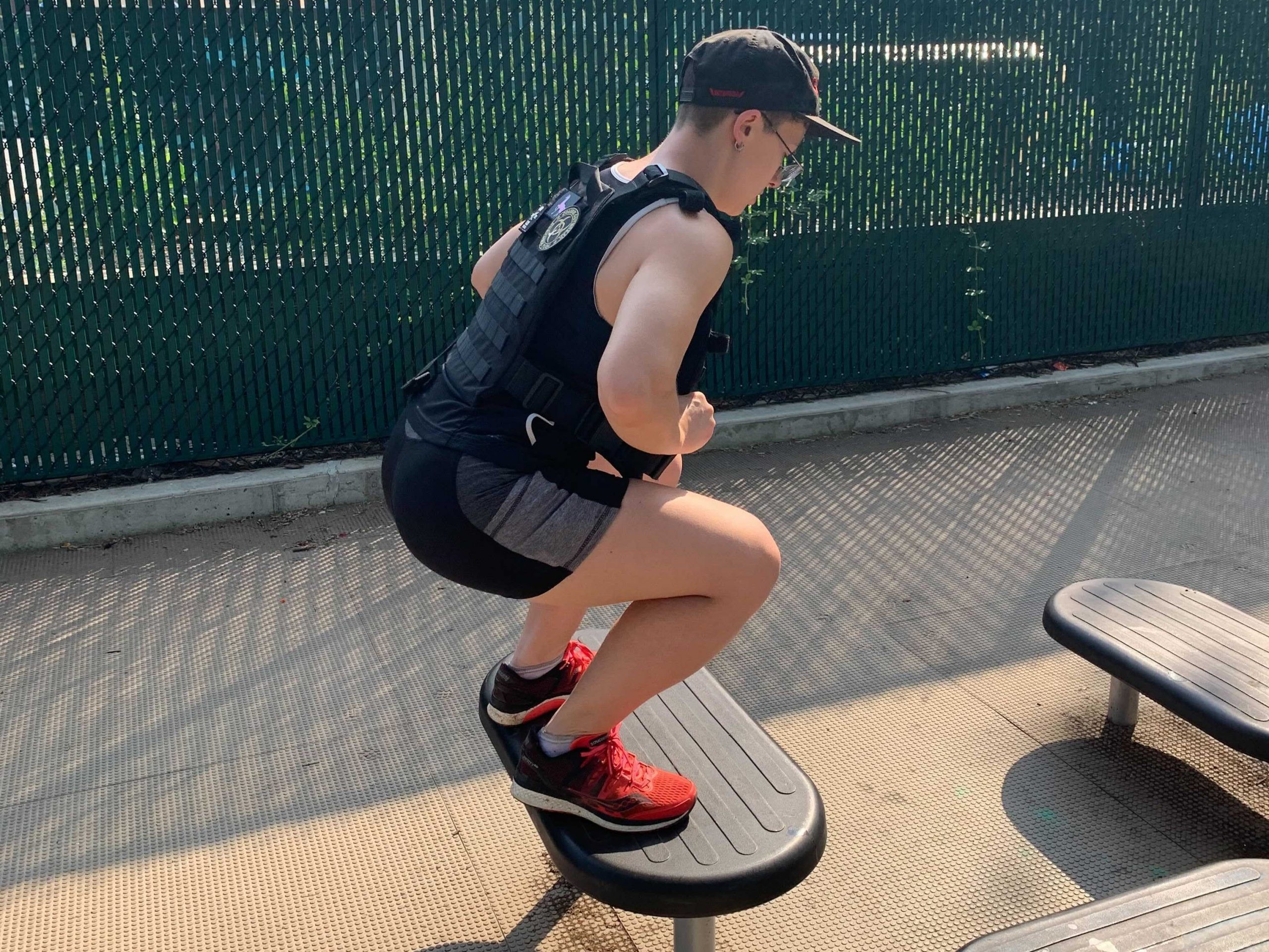 a sweaty fitness reporter in a weighted vest jumping onto a piece playground equipment in a sunny outdoor area next to a green fence