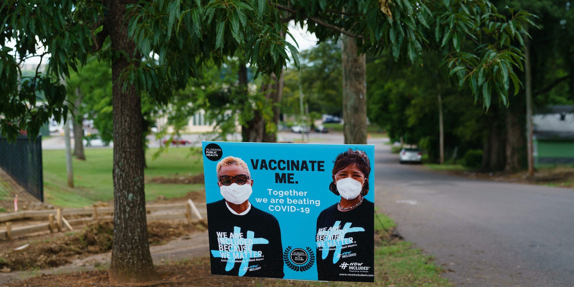 a sign read "vaccinate me" near a road in Alabama. On the sign are two masked people. In the background are trees