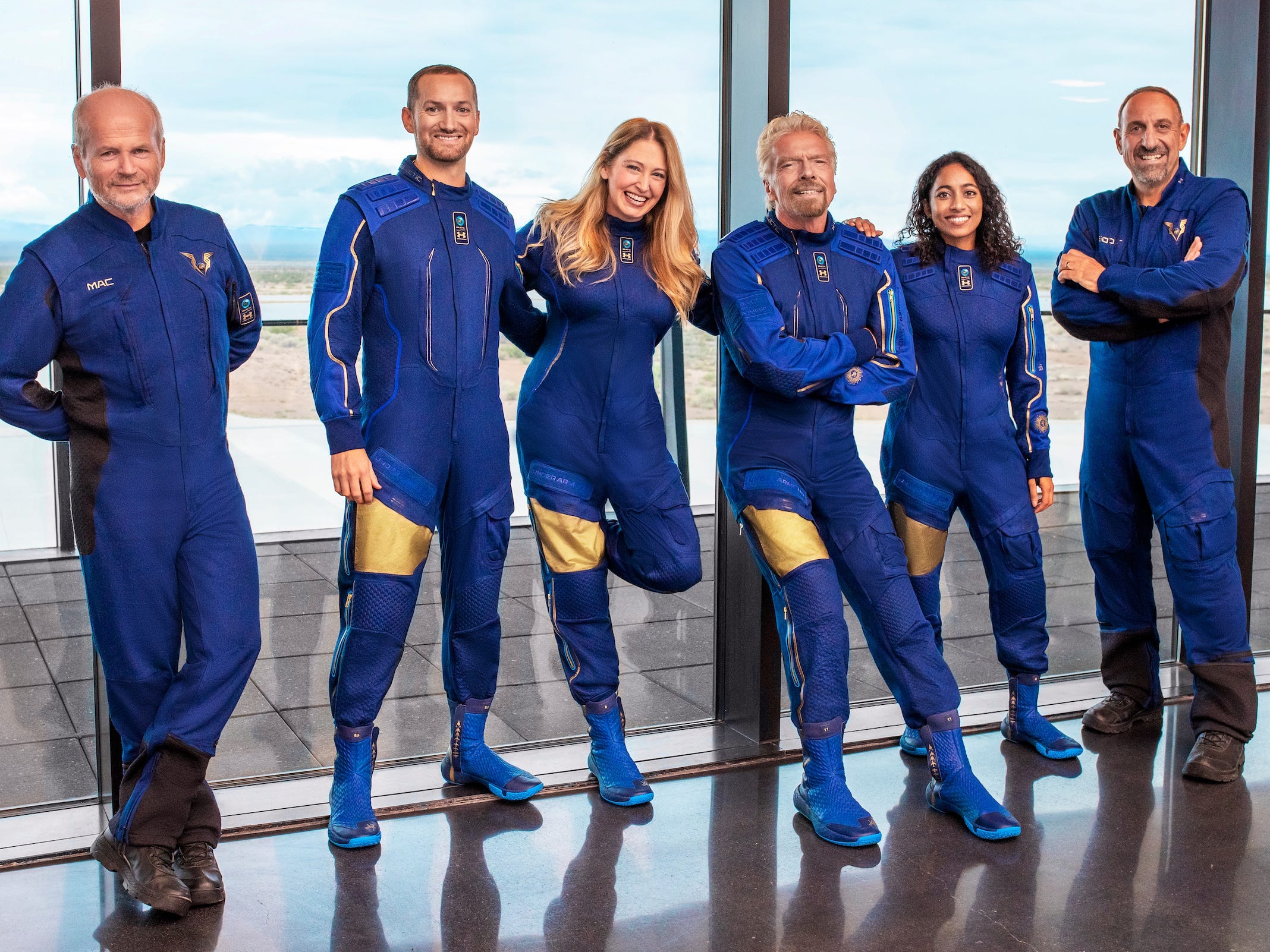 Six people in blue bodysuits - the crew of the VSS Unity - stand in front of a floor-to-ceiling window.