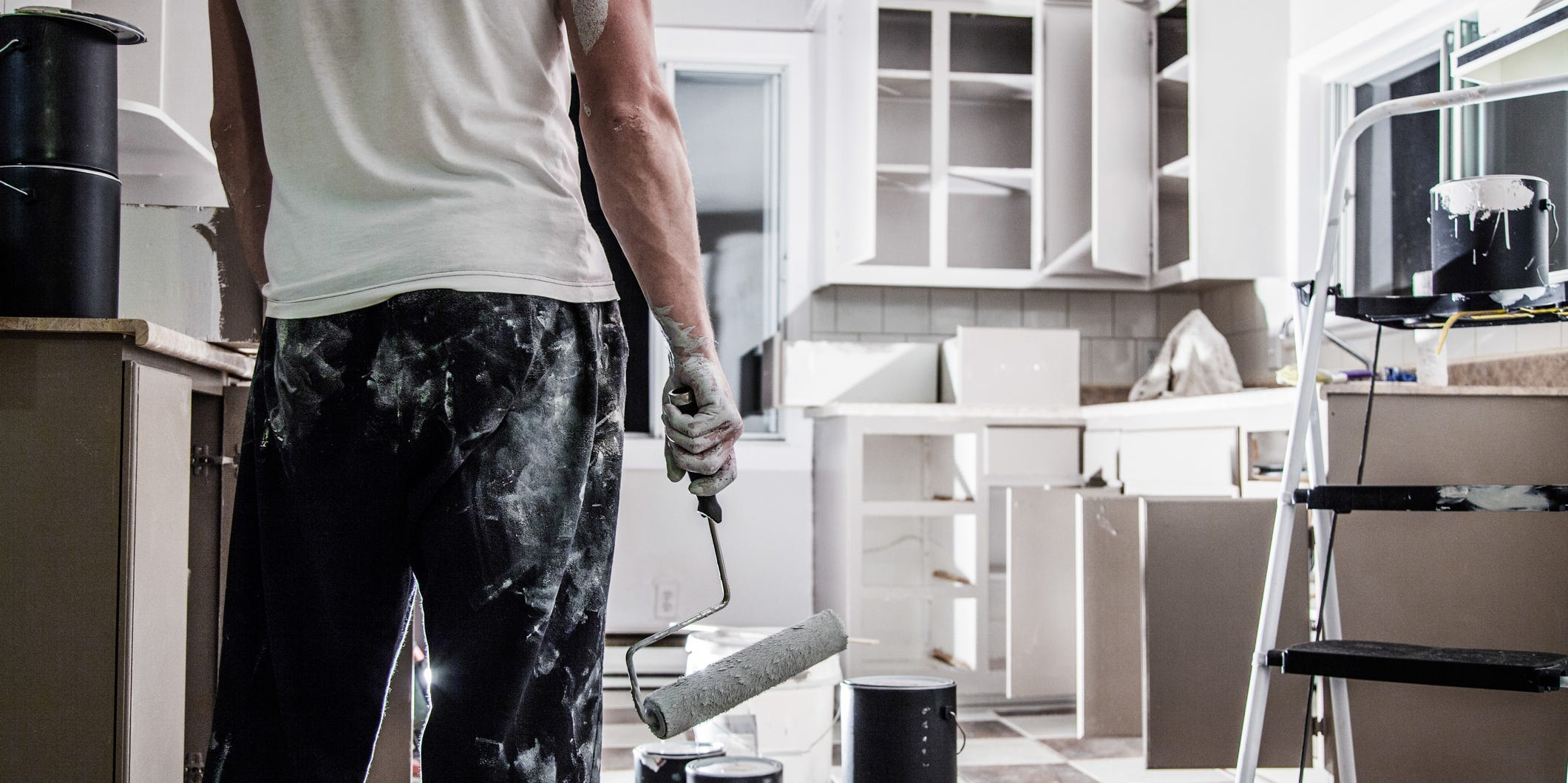 A person covered in paint and holding a paint roller standing back and admiring freshly painted kitchen cabinets