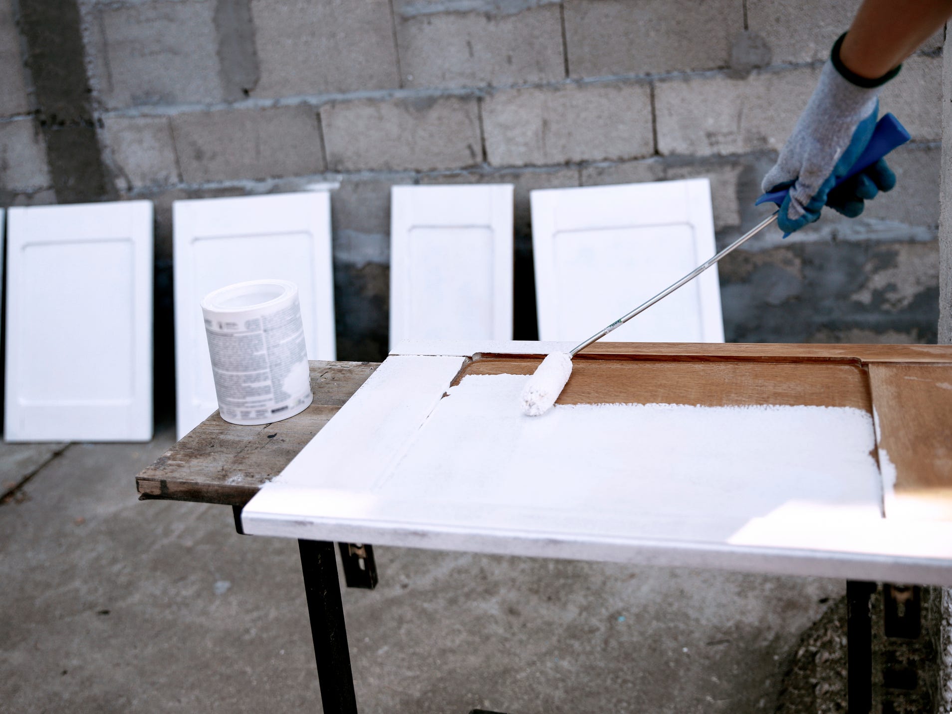 A person painting a wooden kitchen cabinet door white. Other painted cabinet doors are leaned against the wall behind them.