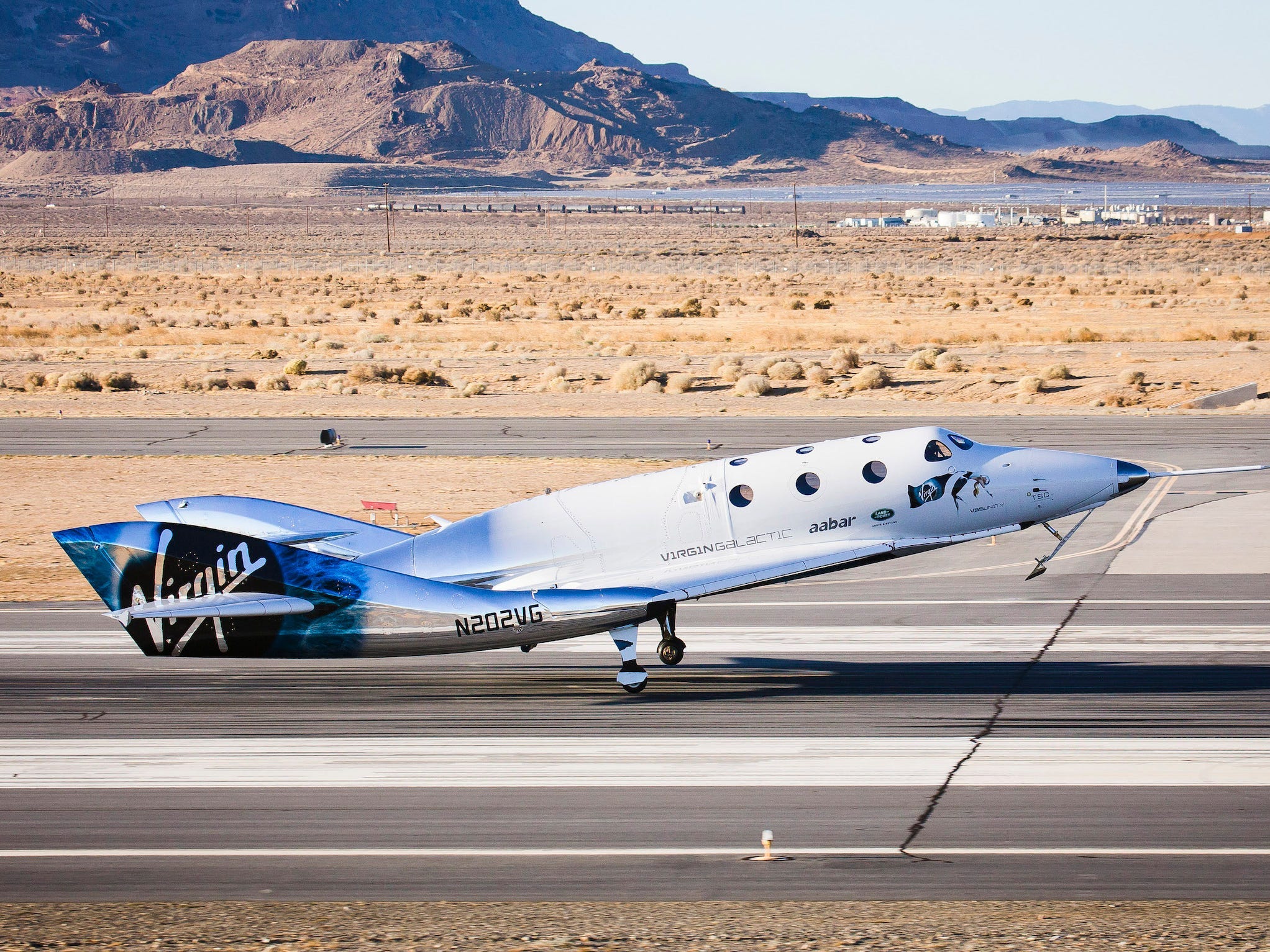 A white-and-silver plane with "Virgin" on the side touching down on a runway, with desert and mountains in the background.