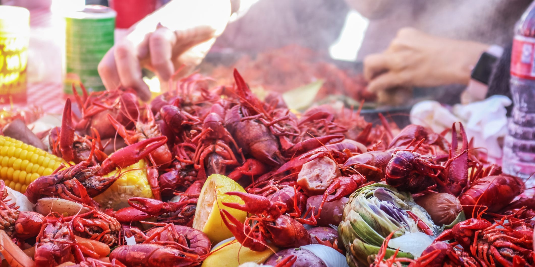 A large pile of steaming boiled crawfish spread on a table with lemon halves, corn on the cob, potatoes, and more interspersed