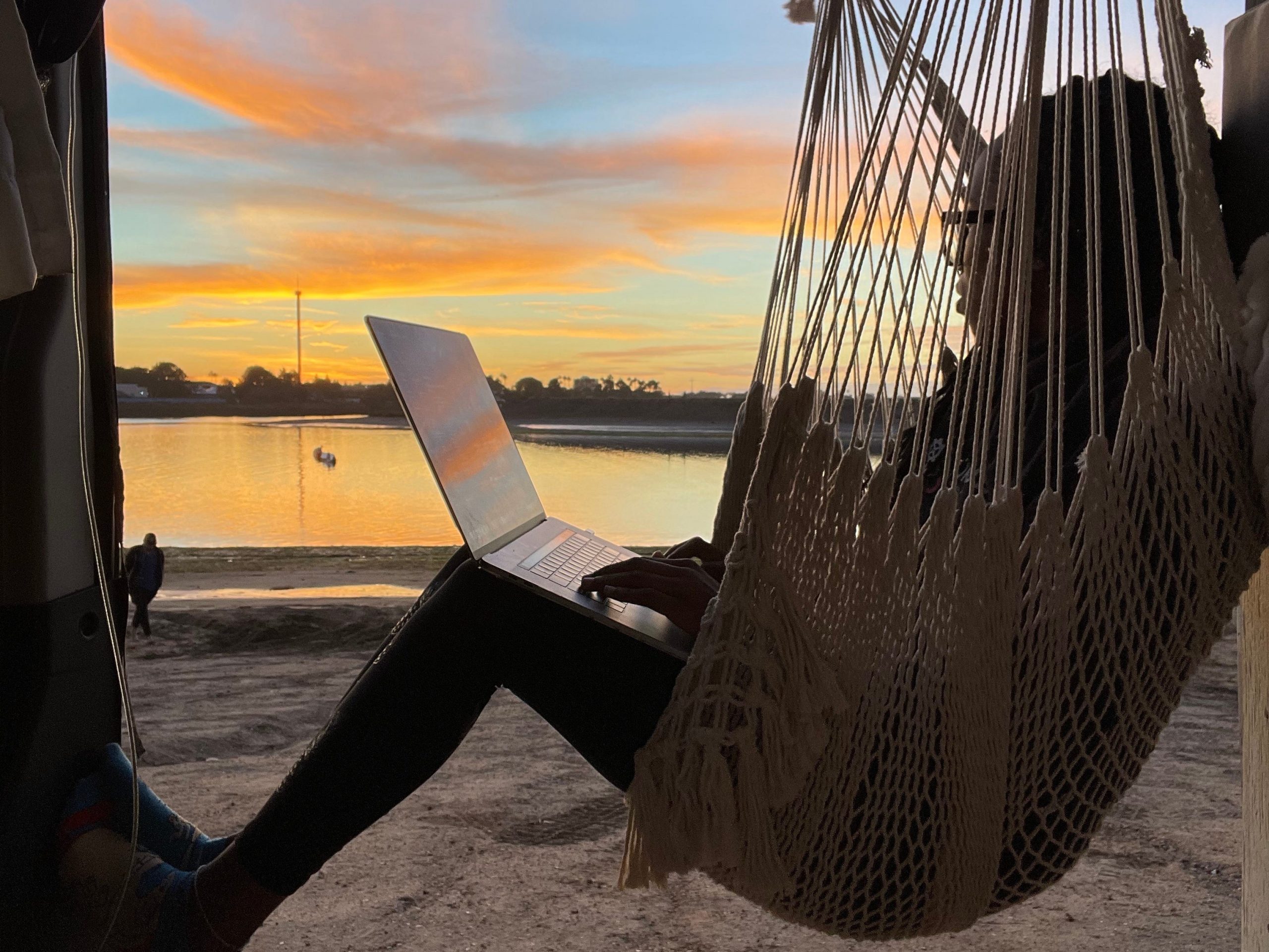 woman sitting in a hammock holding a laptop next to a sunset