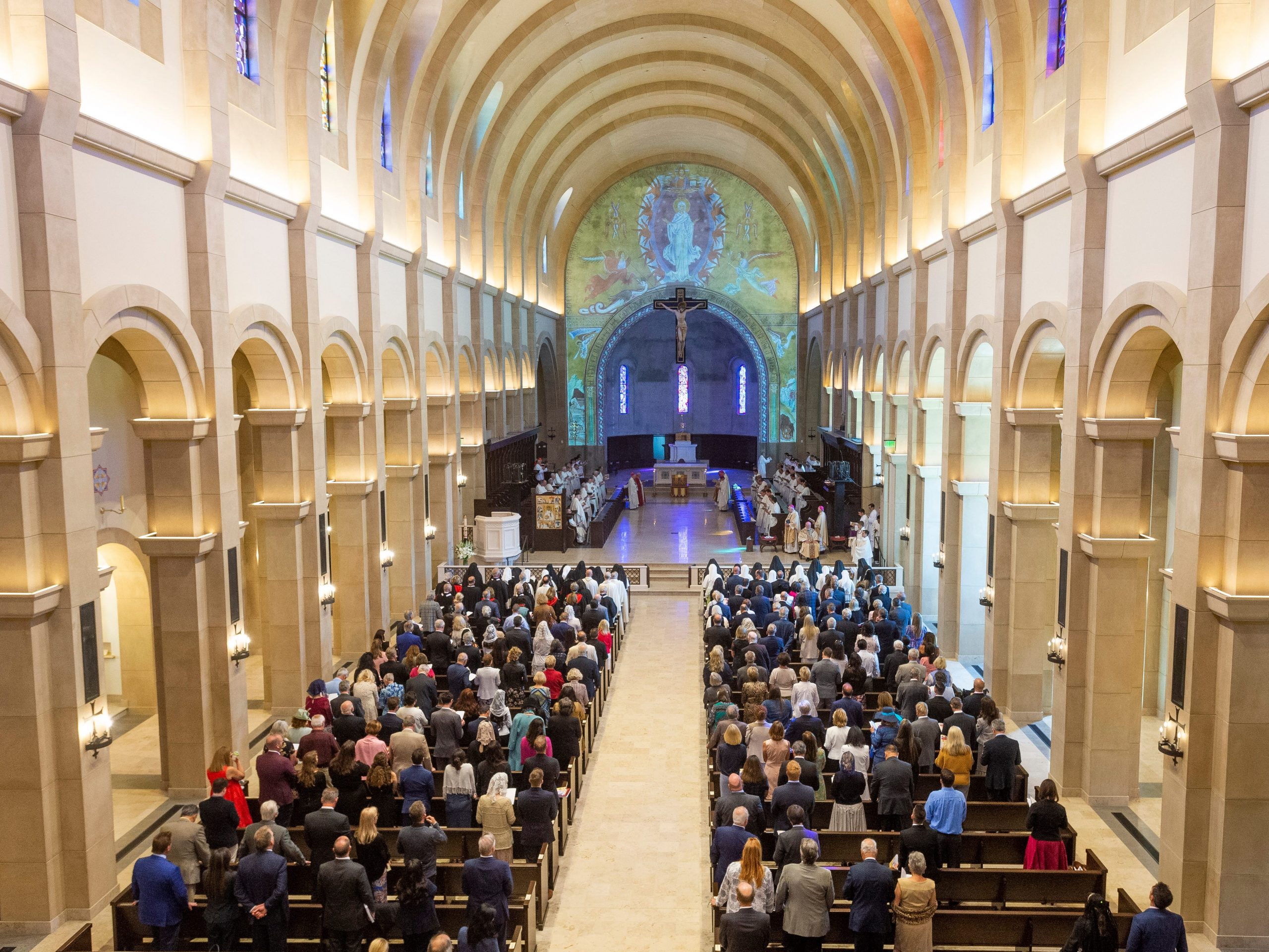 A large church with an arched ceiling and alter lit up by colored lights and pews packed with people.