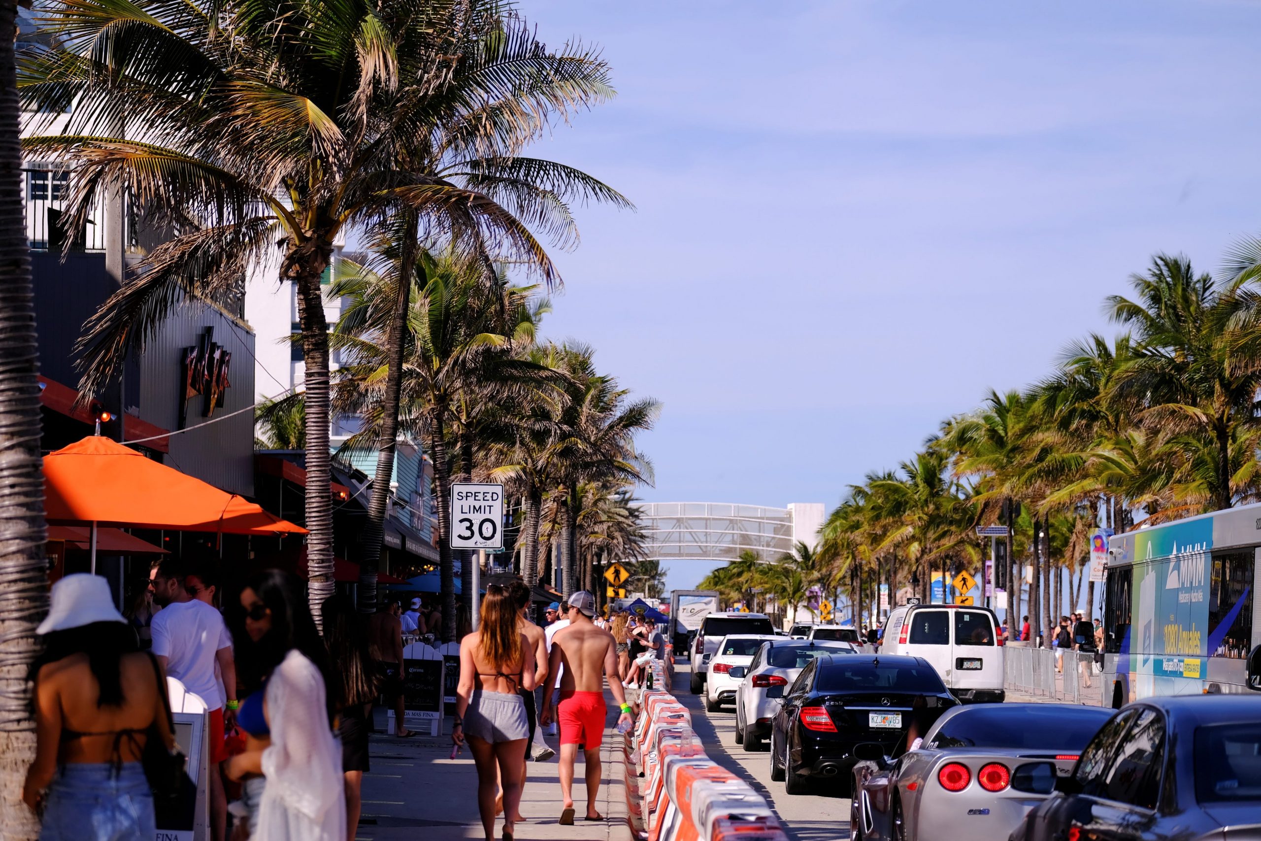Beachgoers in bathing suits walk alongside a row of stopped traffic in sunny Fort Lauderdale