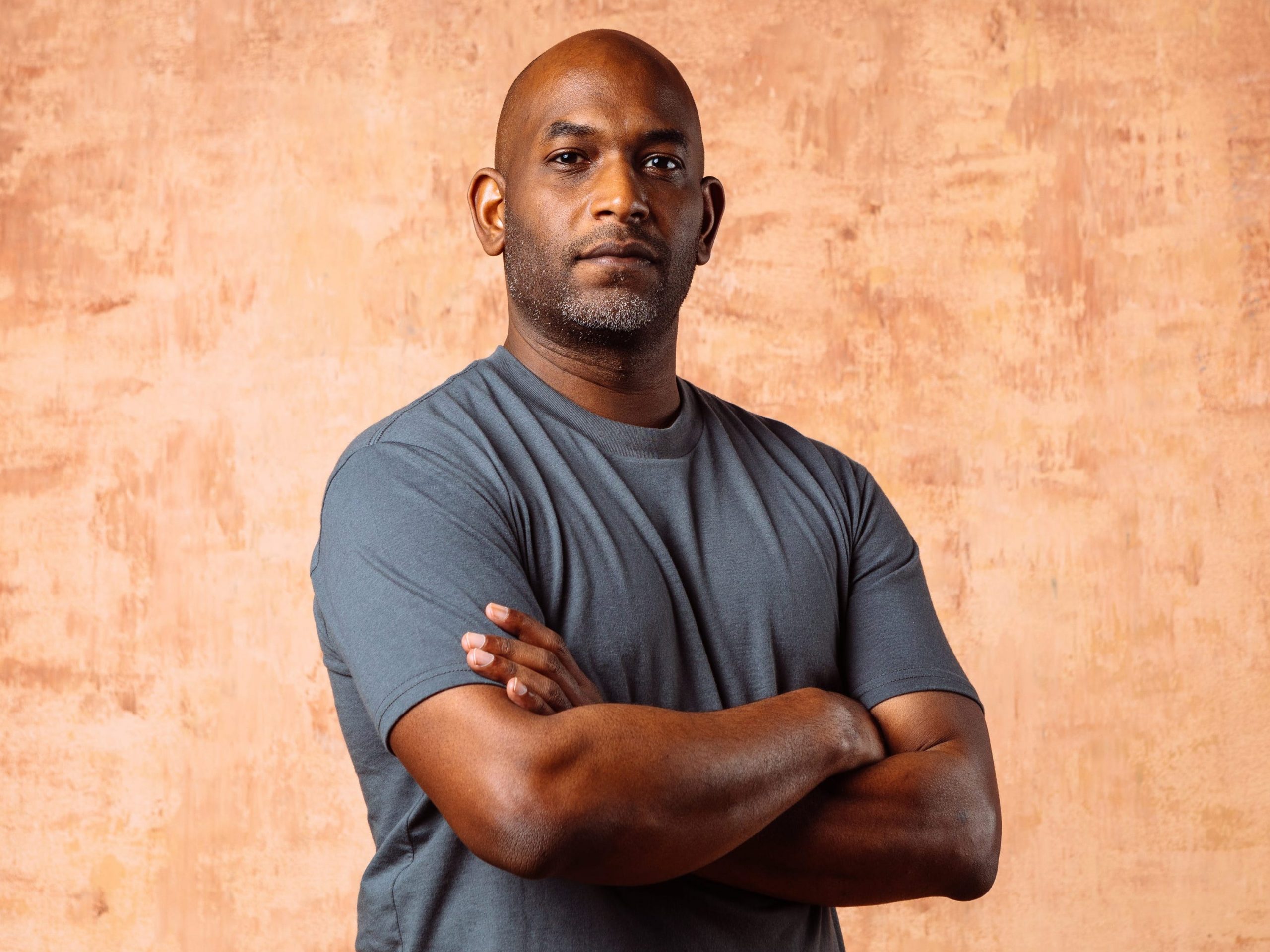 Kelsey Hightower, principle engineer at Google and self-taught developer. His arms are crossed and he's wearing a grey shirt in front of a textured brown background.