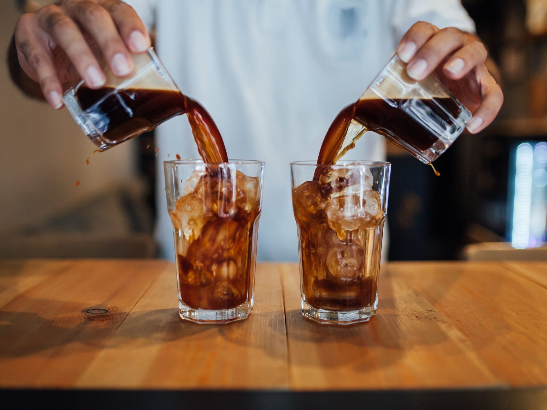 A pair of hands pouring two small glasses of cold brew into two larger glasses full of ice