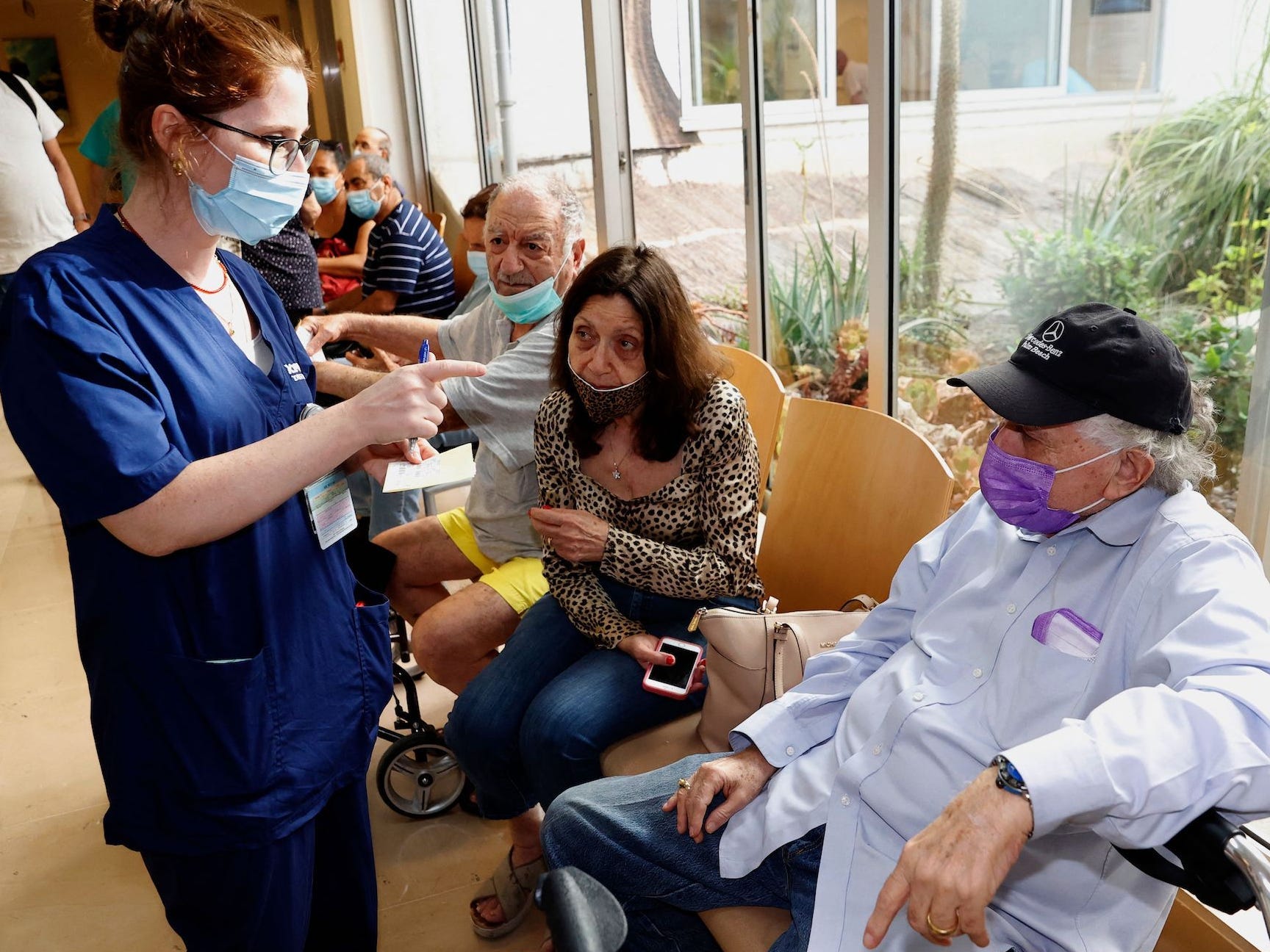 A woman in blue scrubs wearing a mask talks an older person wearing a black cap wearing a purple mask and white shirt while others wait sitting on chairs in the background.