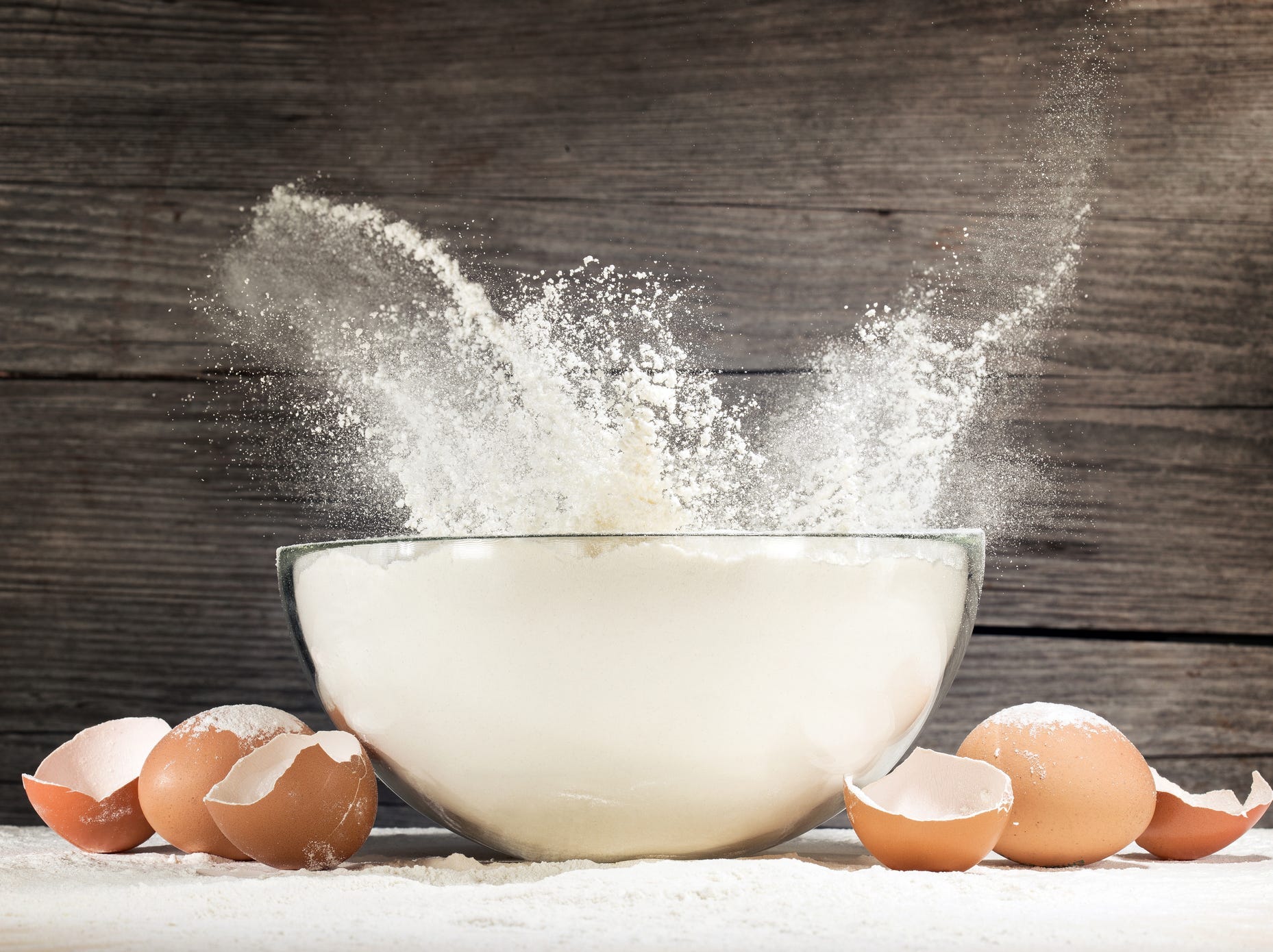 Flour splashing out of a glass bowl surrounded by broken eggshells