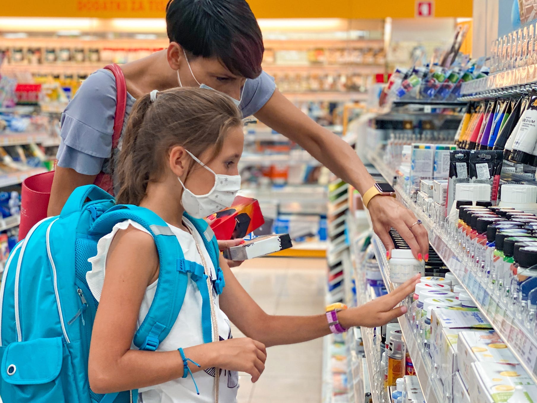 Mother and daughter wearing face masks and looking at school supplies in a store. The daughter is wearing a blue backpack.