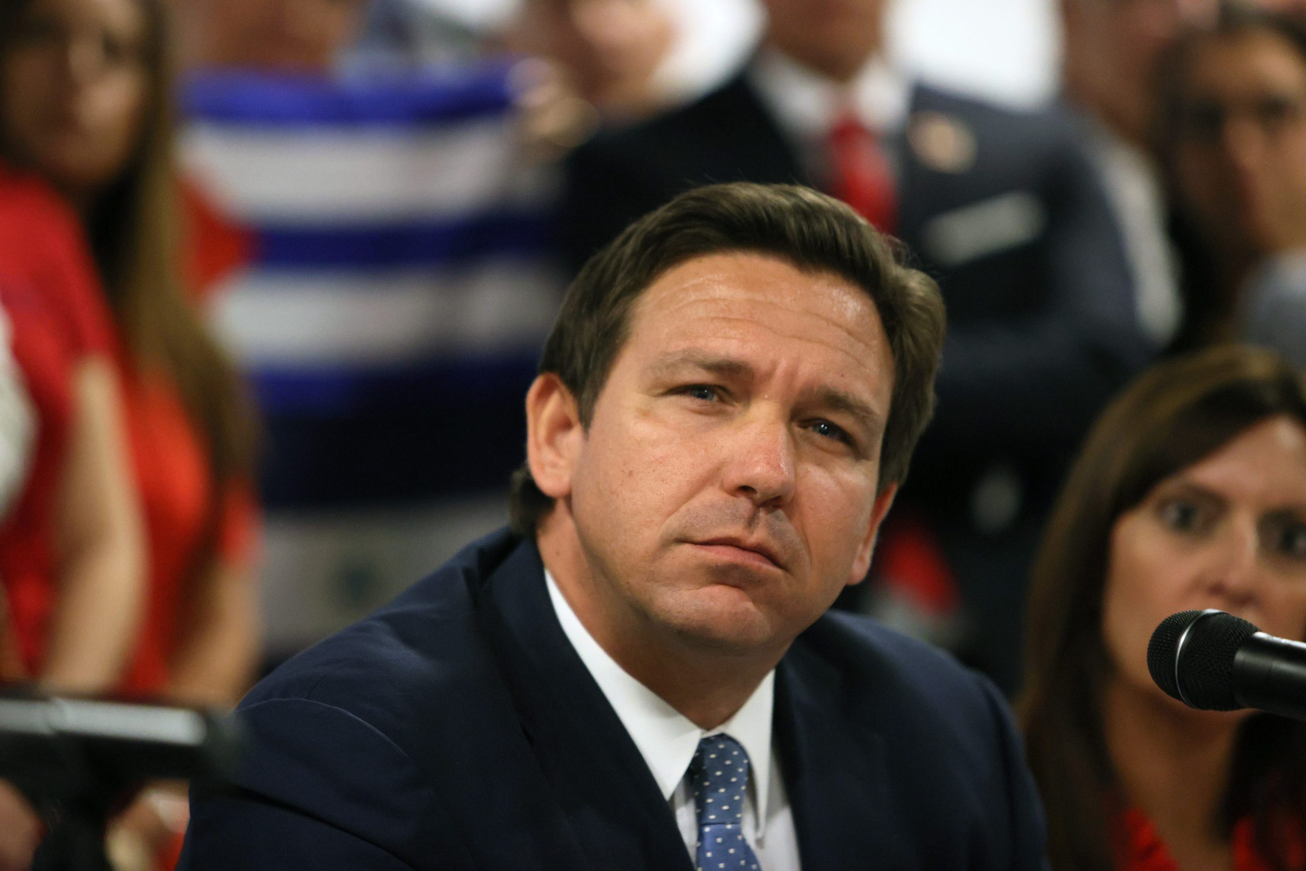 Florida Governor Ron DeSantis wears a black suit and blue tie and looks into the distance.