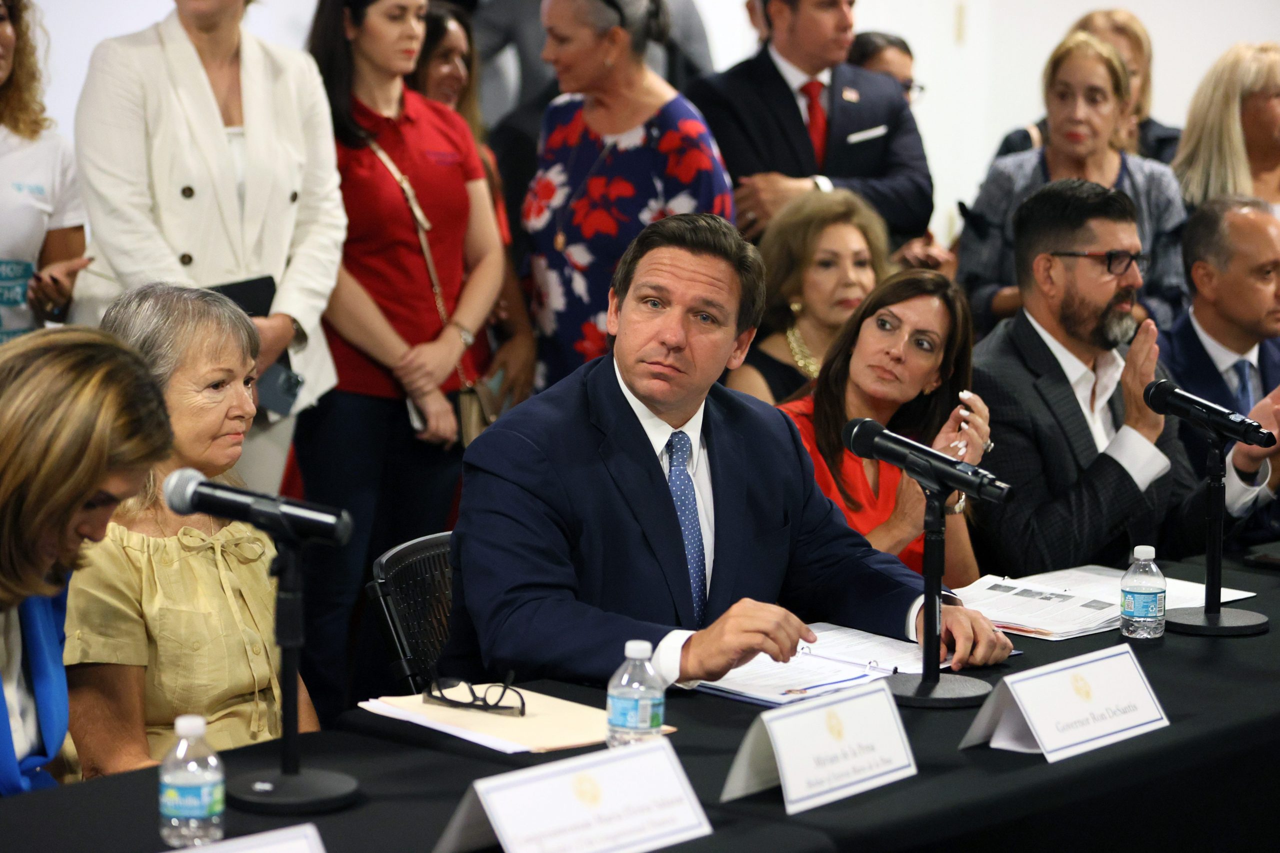 Florida Governor Ron DeSantis sits at a table wearing a black suit with a blue tie surrounded by people.