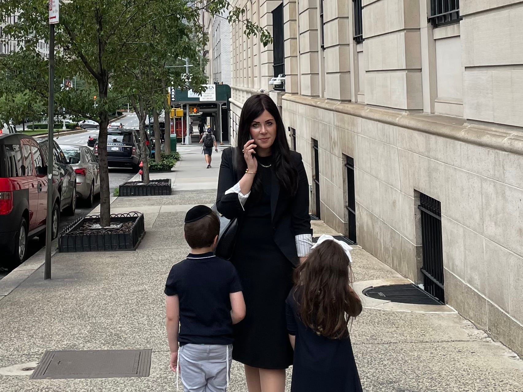 A woman on the phone stands in the middle of the sidewalk with her two kids facing towards her