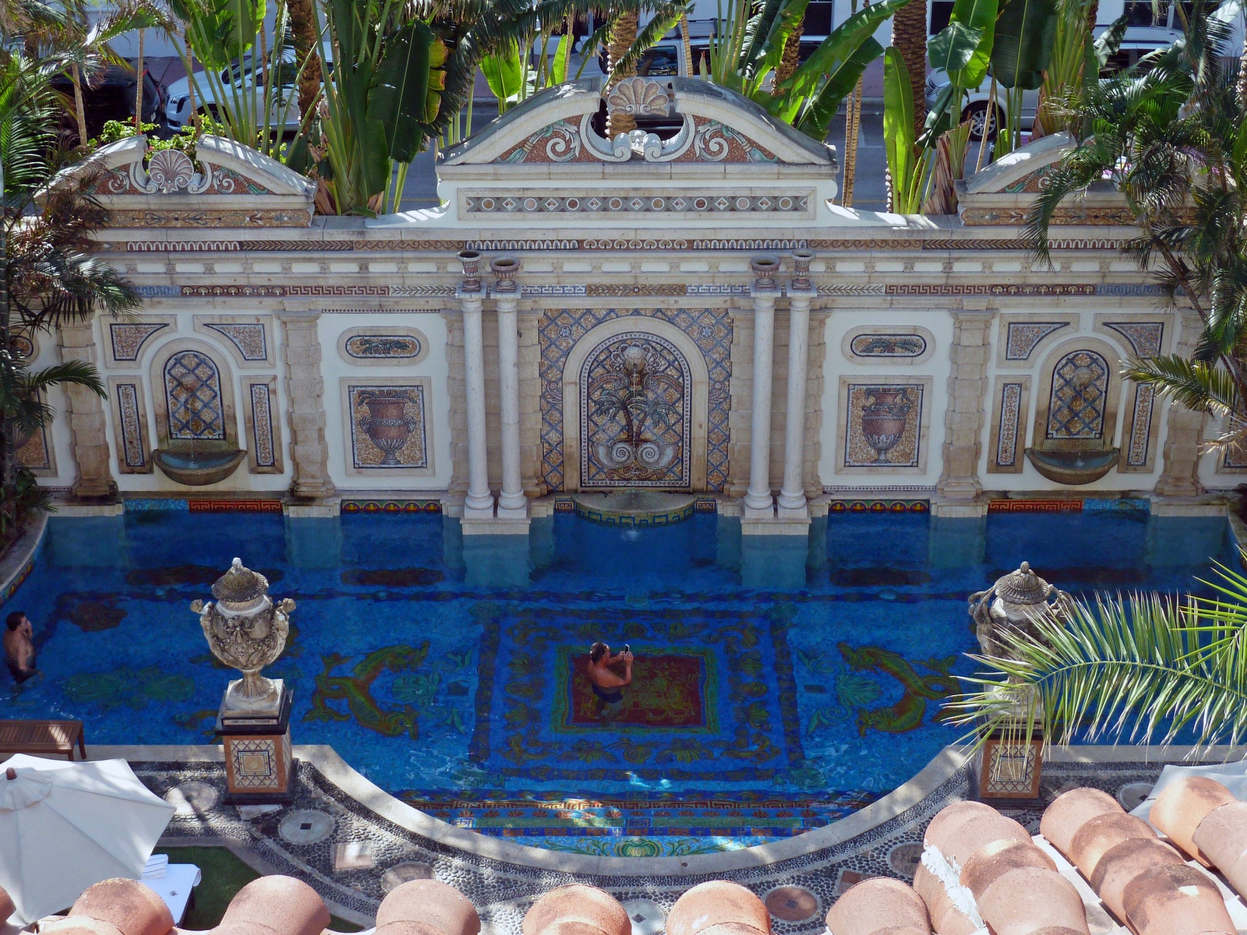 Two men swim in an elaborately designed swimming pool next to a wall of stone columns and a fountain at the former Versace mansion.