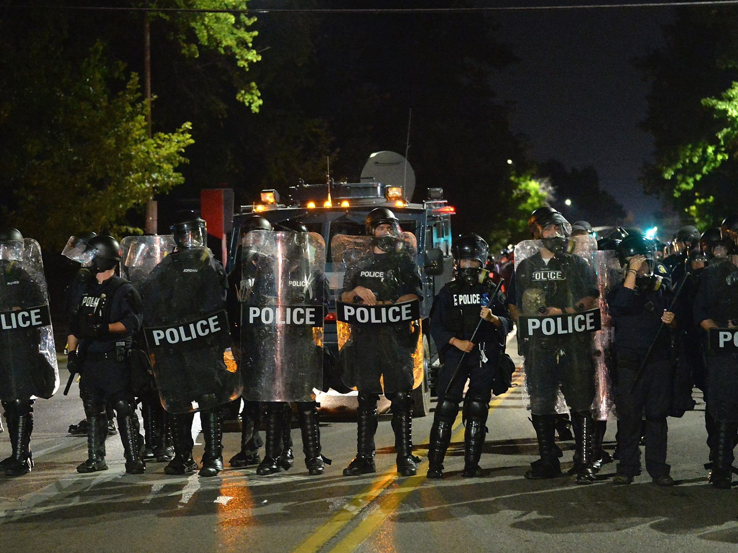 A line of police officers in riot gear stand in a line across a street.