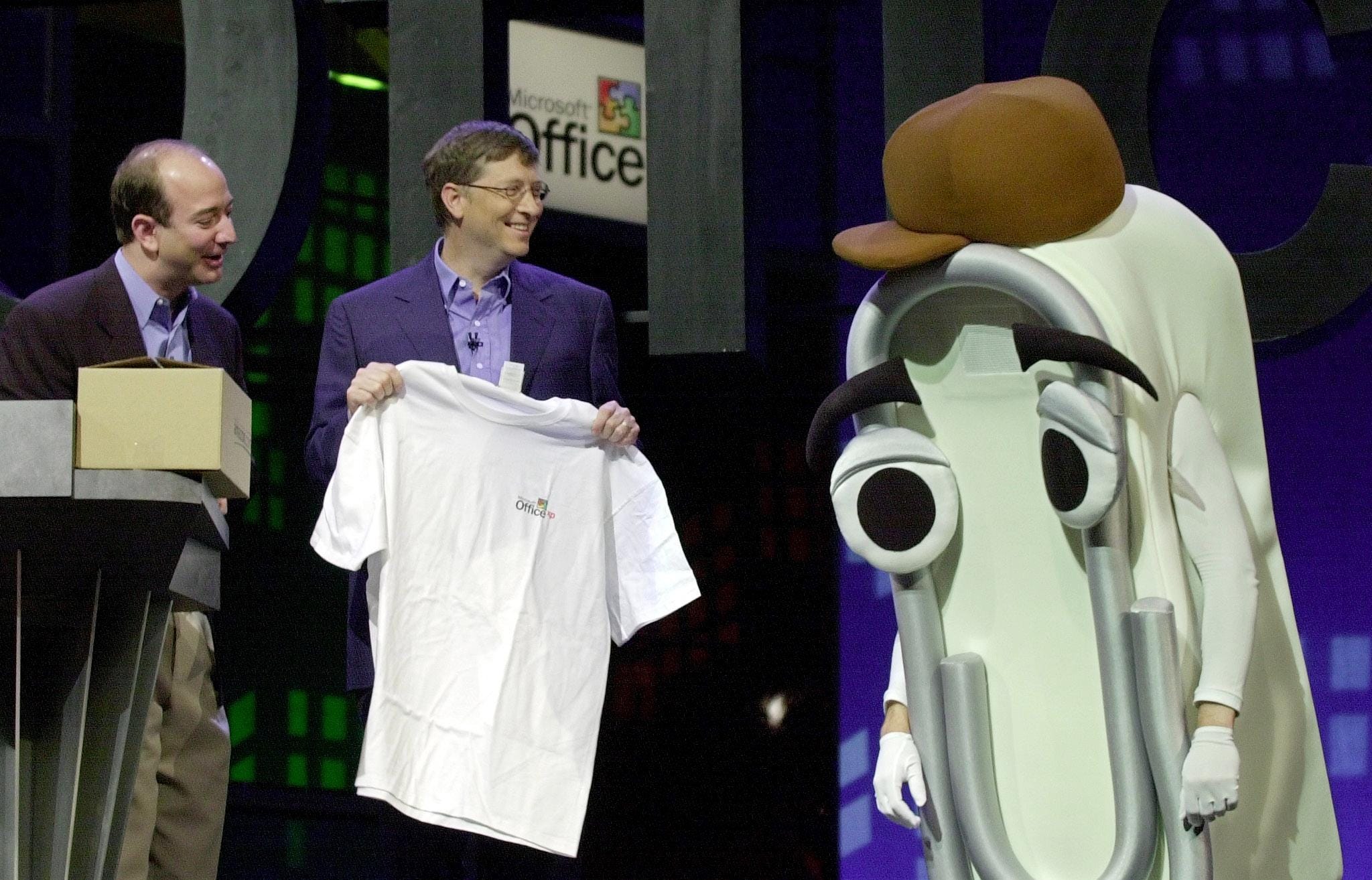 Former Microsoft CEO Bill Gates and former Amazon CEO Jeff Bezos hand a white t-shirt to someone dressed in a cartoon paperclip costume.