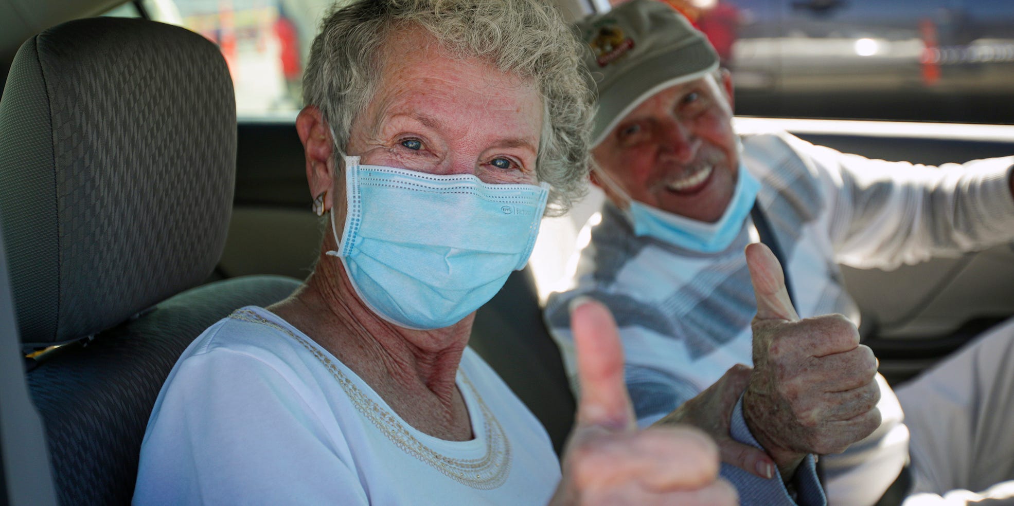 Two older people smile and hold their thumbs up while sitting in the front of a car