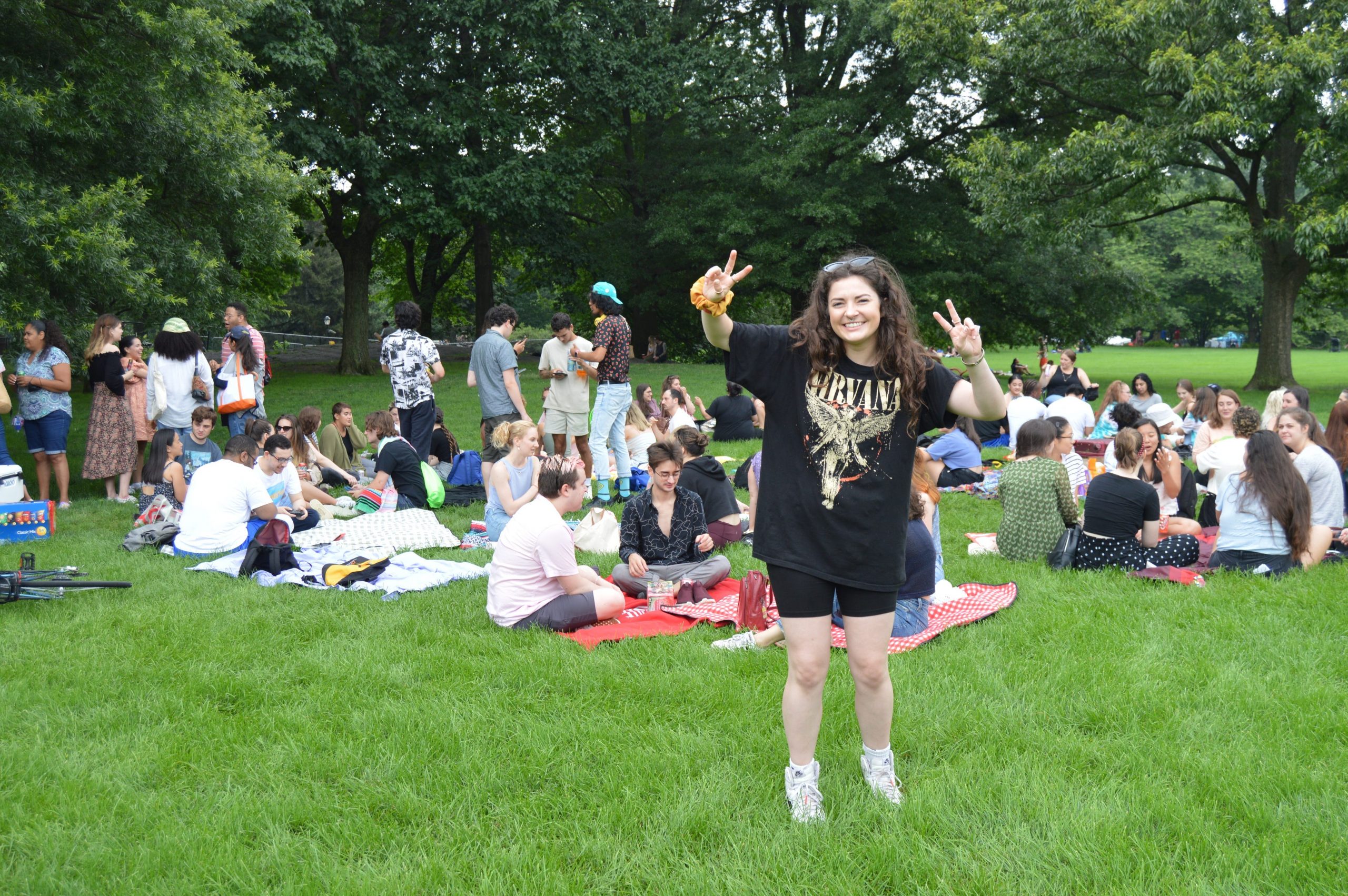 A girl with brown hair wears a black T-shirt and black shorts in front of a large group sitting in a park. She is smiling and holding her hands in two peace signs.