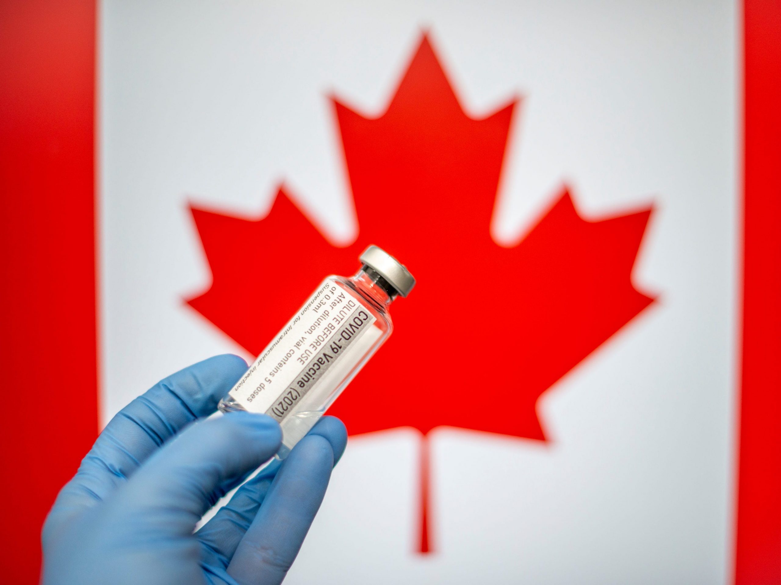 A gloved hand holding a vial of COVID-19 vaccine in front of a Canadian flag