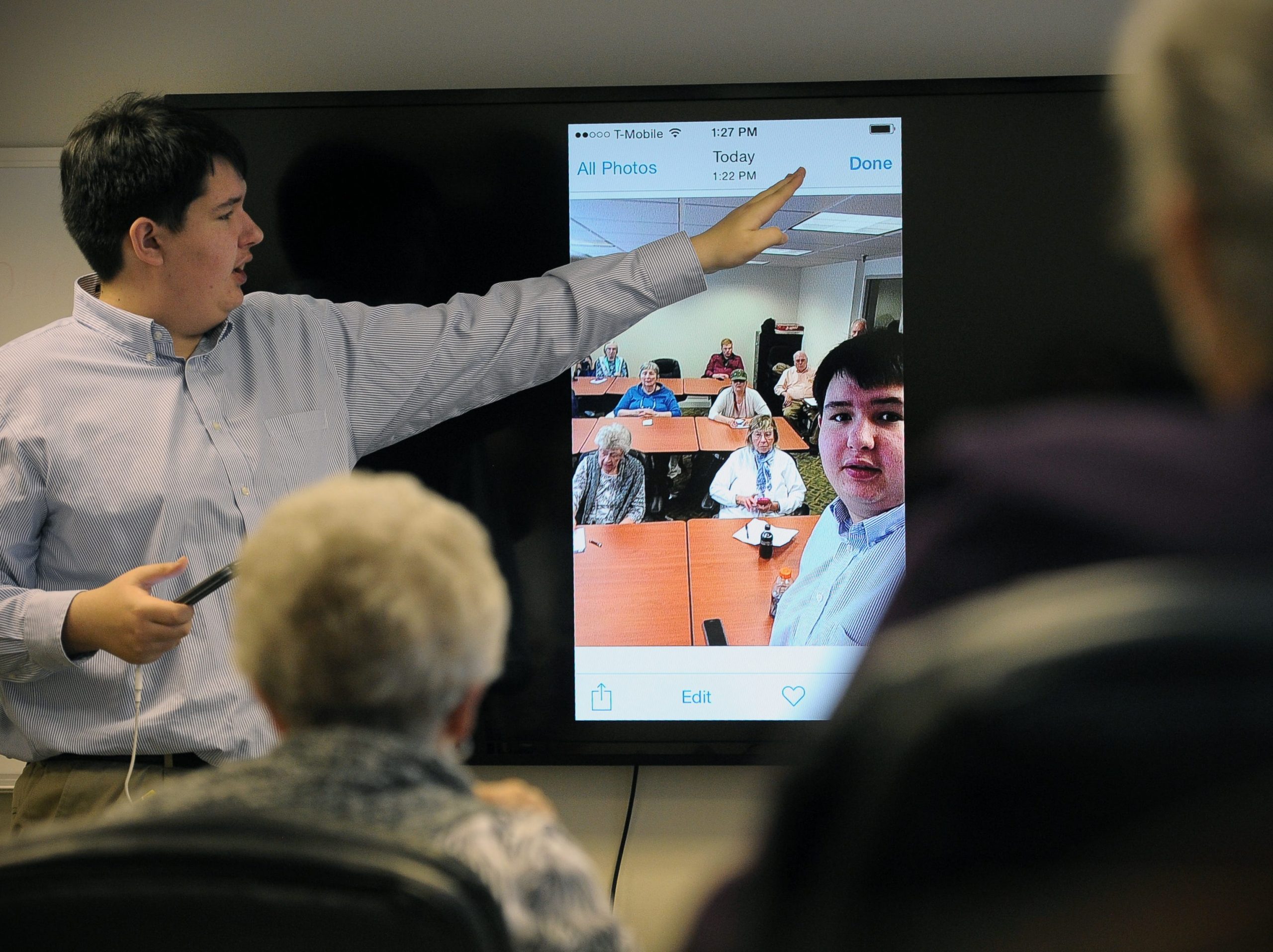 A 16-year-old boy in a button up gestures to a phone screen portrayed on a television in front of a room of old people.