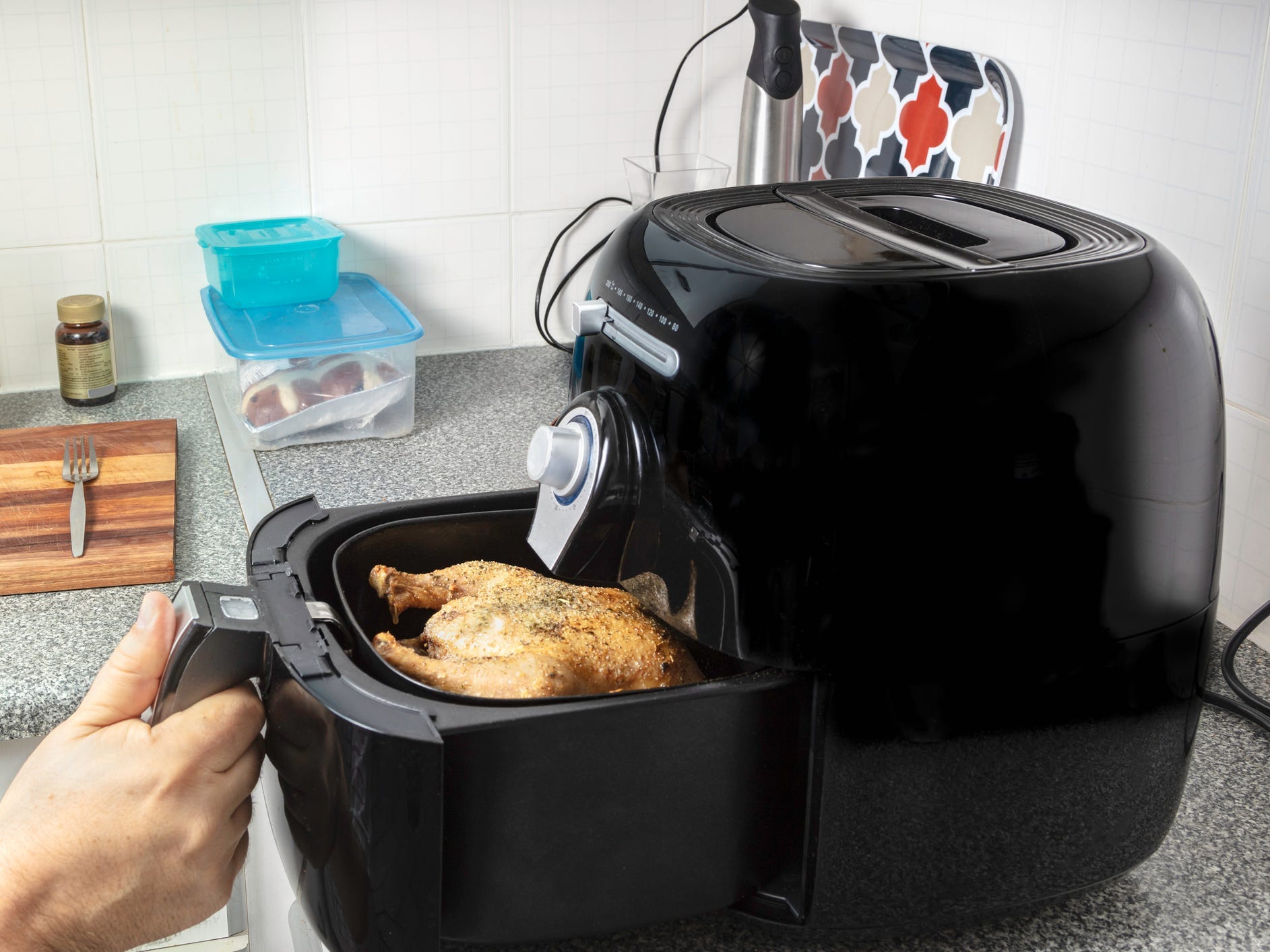 A hand pulling out the basket of an air fryer revealing a whole chicken