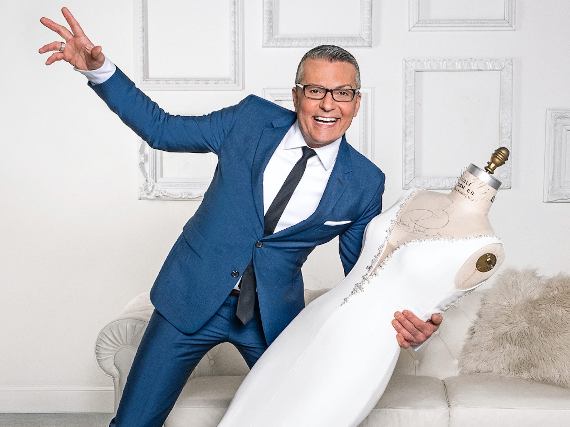 Randy Fenoli holds a mannequin dressed in a white wedding dress in front of a white wall.