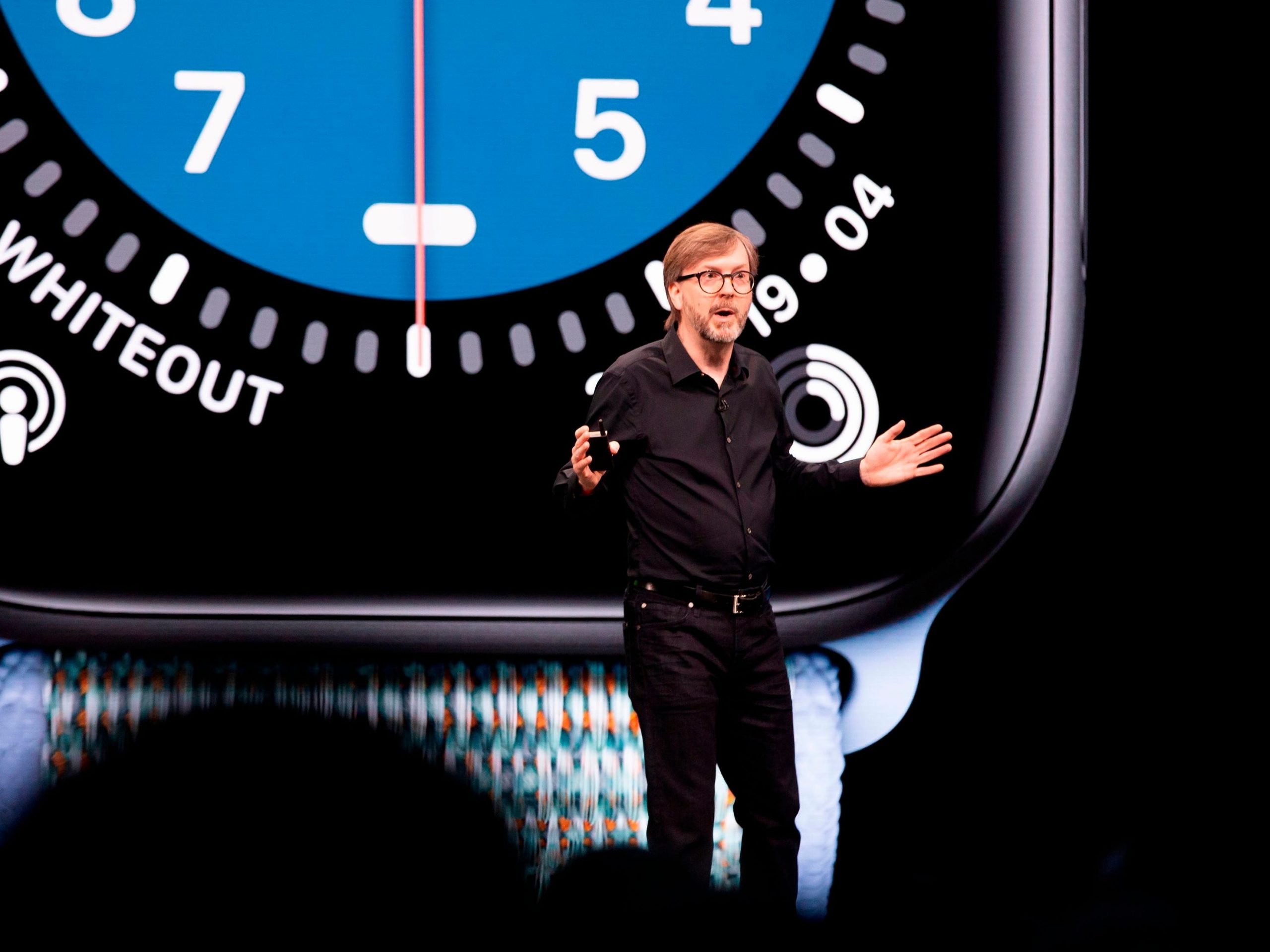 Kevin Lynch gestures and speaks on a stage in front of a projection of an Apple Watch.