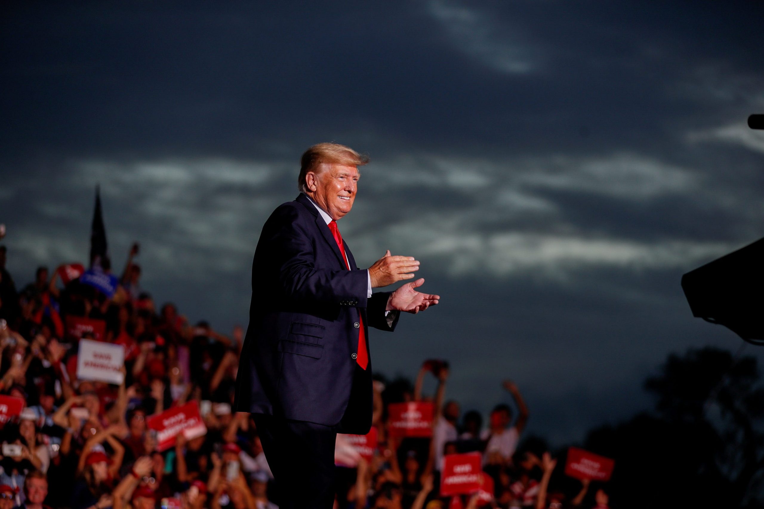 Former President Donald Trump in a red tie and blue suit claps his hands in front of a crowd and a stormy sky