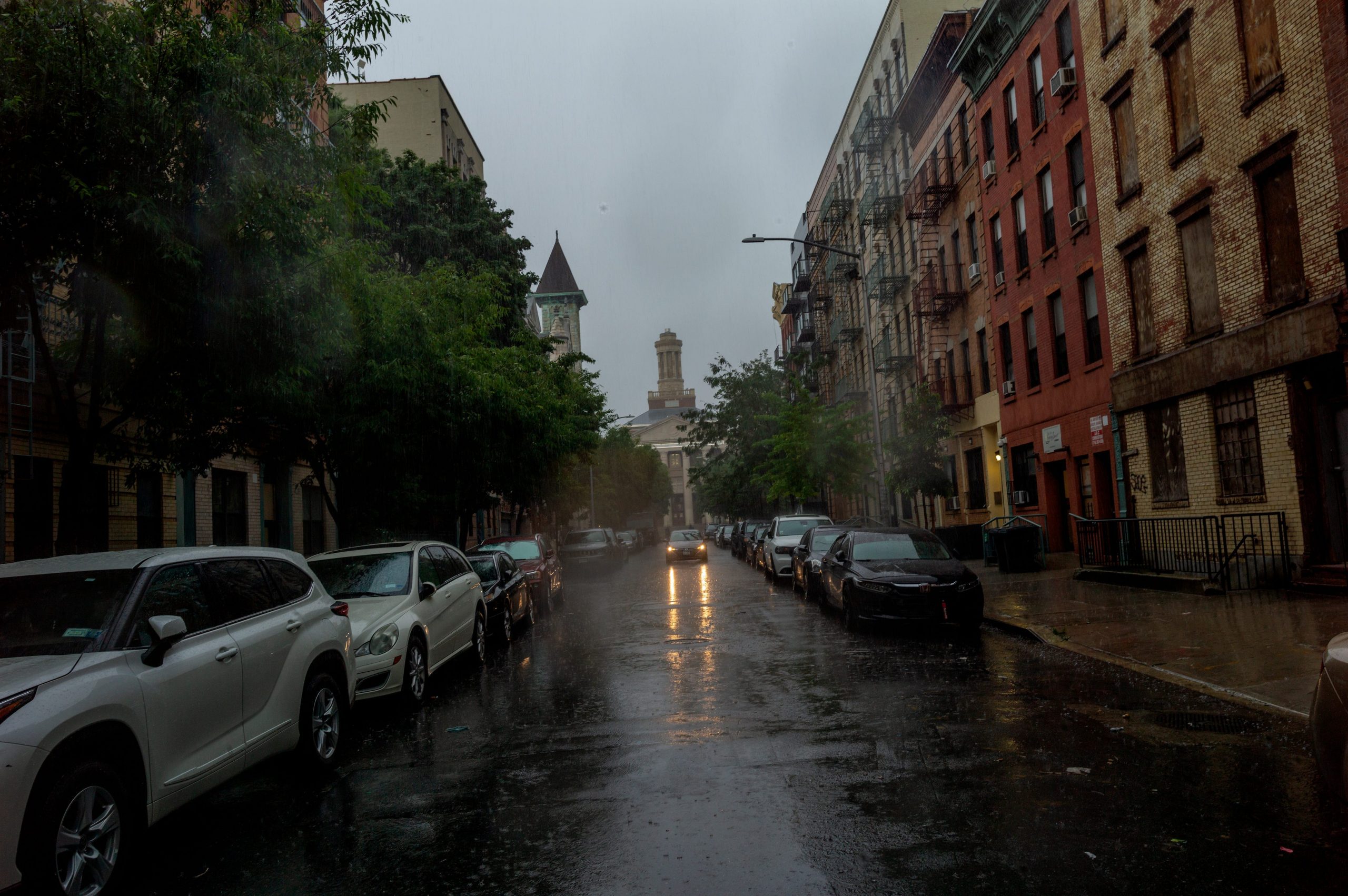 A rainy view of a New York City block with cars parked on either side of the road