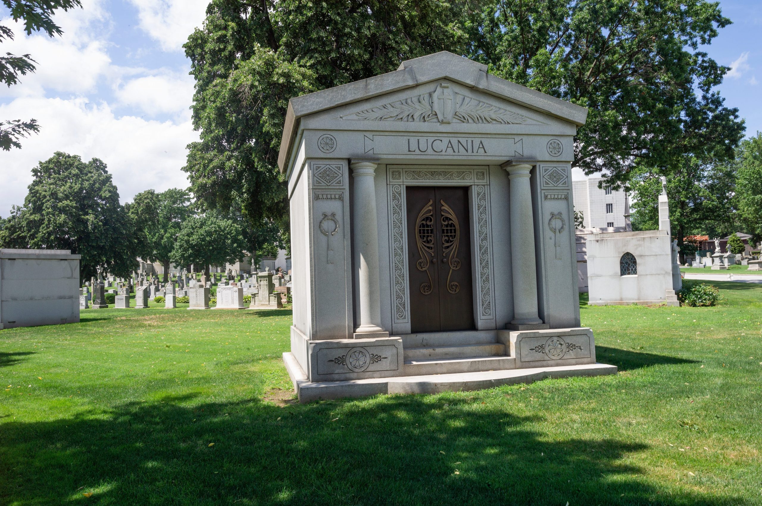 A small mausoleum is seen in a grassy cemetery, with smaller grave stones in behind it.