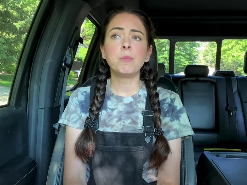 youtuber nikki phillippi looking at the camera and sitting in a car with a forest in the background
