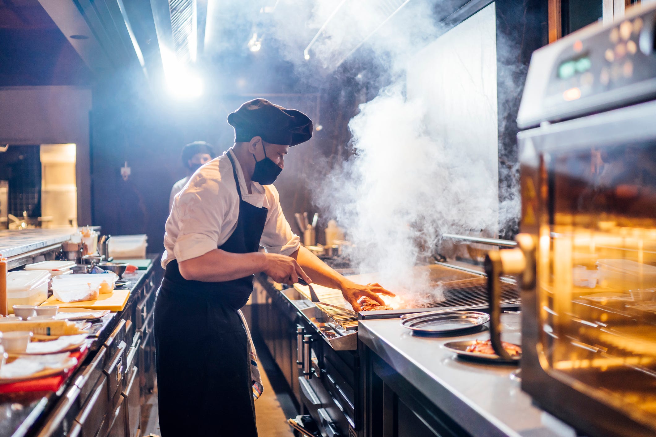A chef in a restaurant kitchen wears a black face mask while cooking.