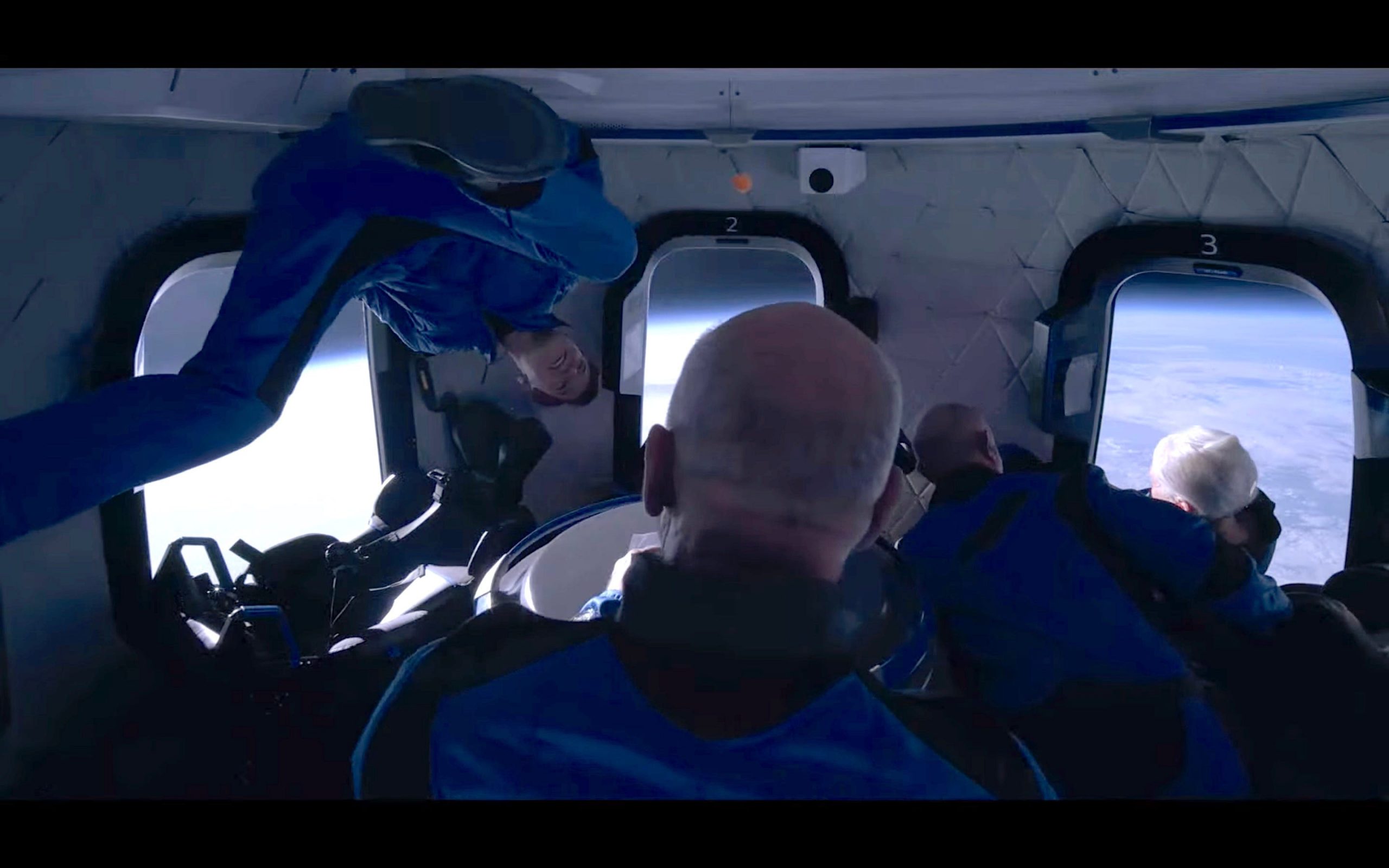 jeff bezos and three other passengers in jumpsuits float around spaceship cabin earth in background