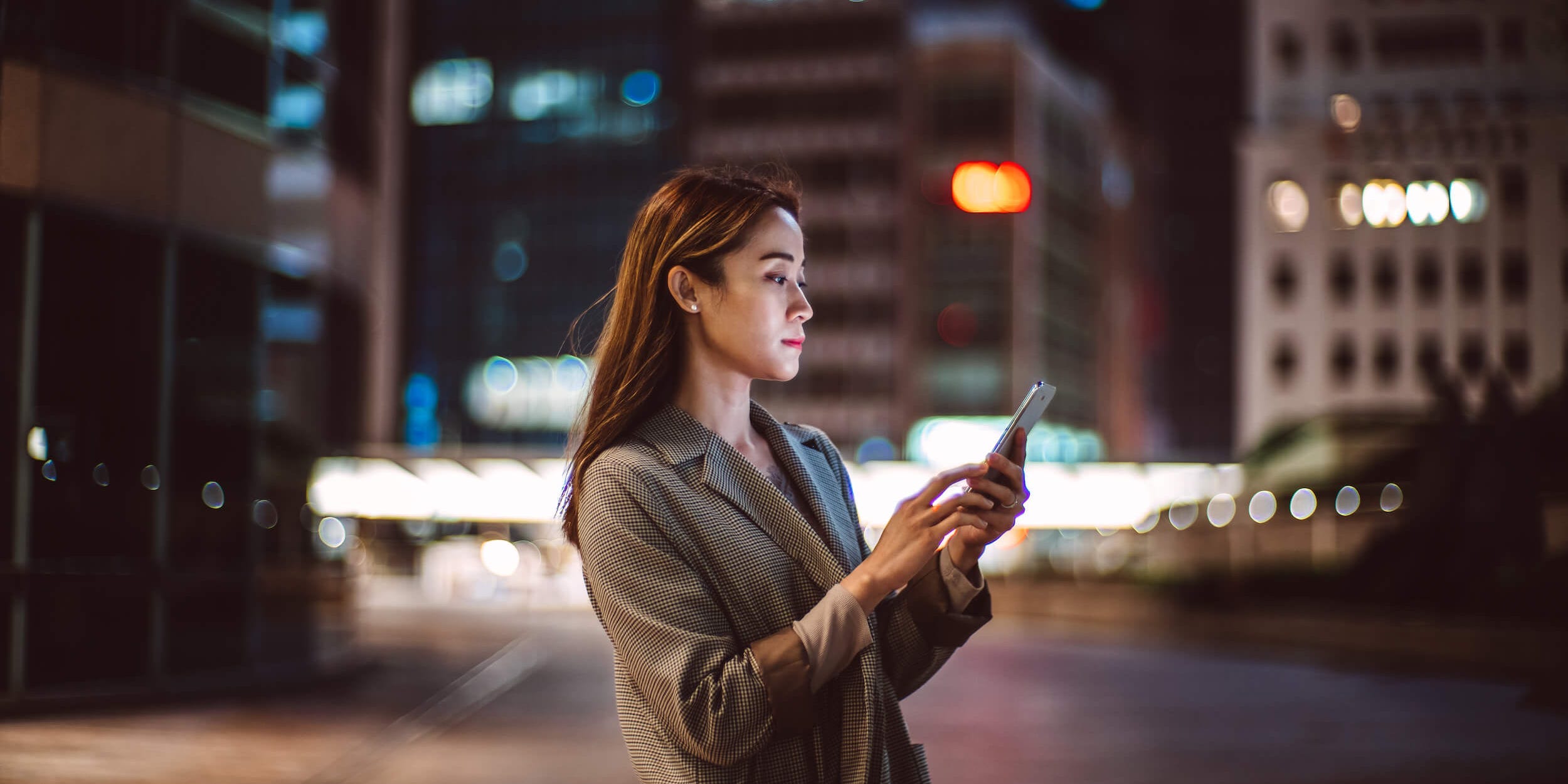 Young Asian woman using a smartphone in downtown district, against illuminated city buildings.