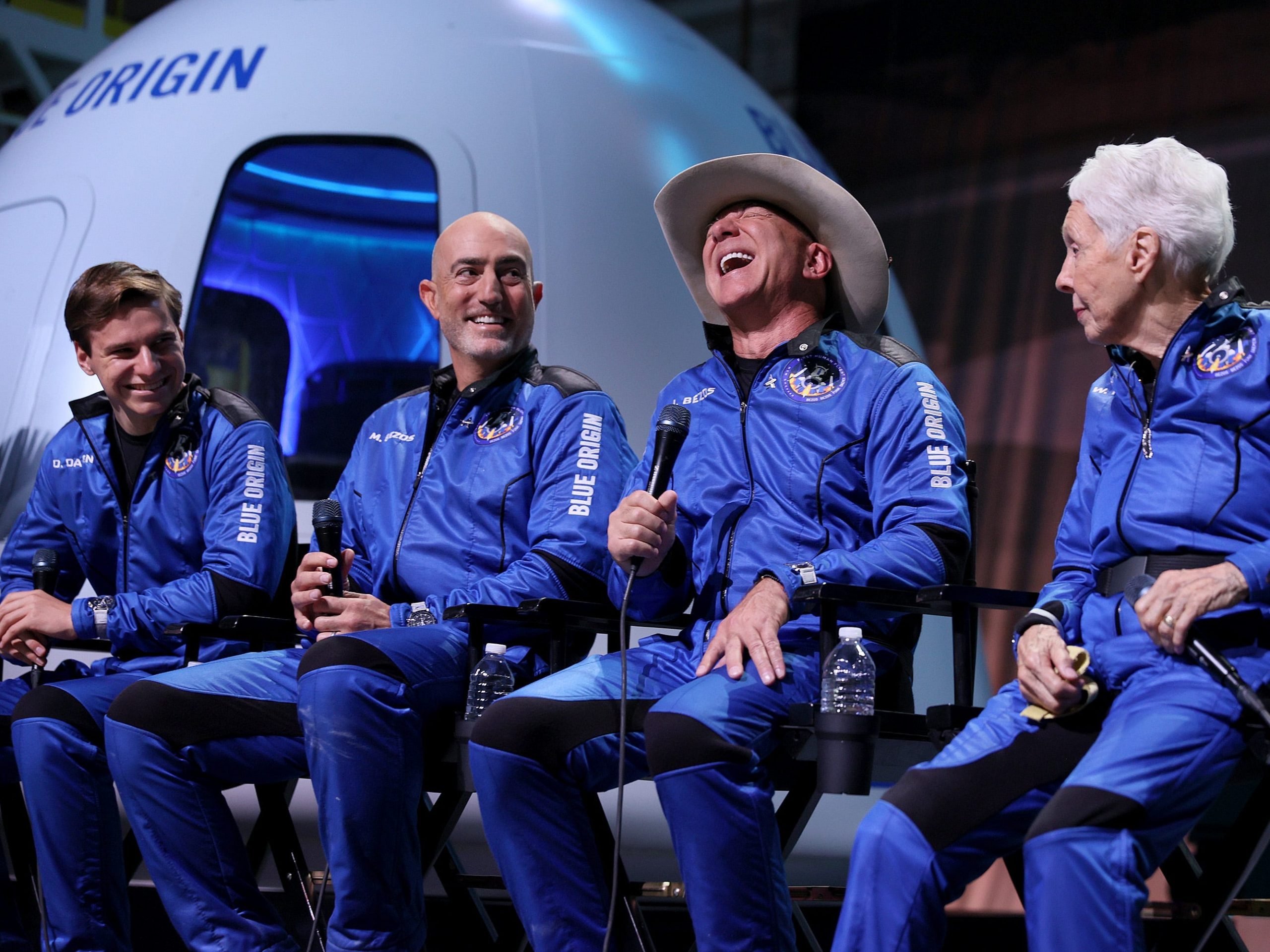 Blue Origin’s New Shepard crew is seen in blue flight outfits sitting in front of the shuttle's capsule.ª