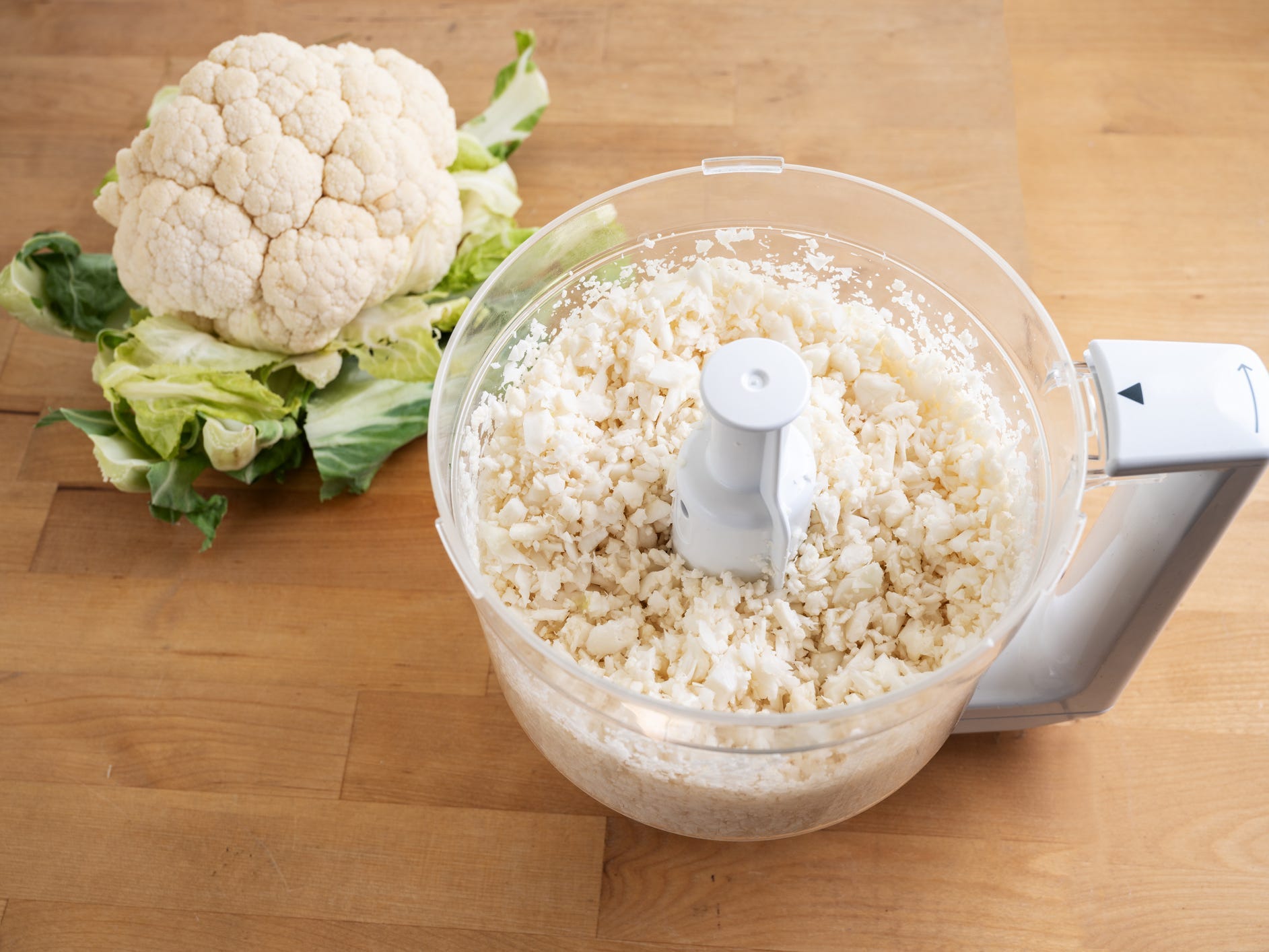 A head of cauliflower next to the bowl of a food processor full of freshly made cauliflower rice