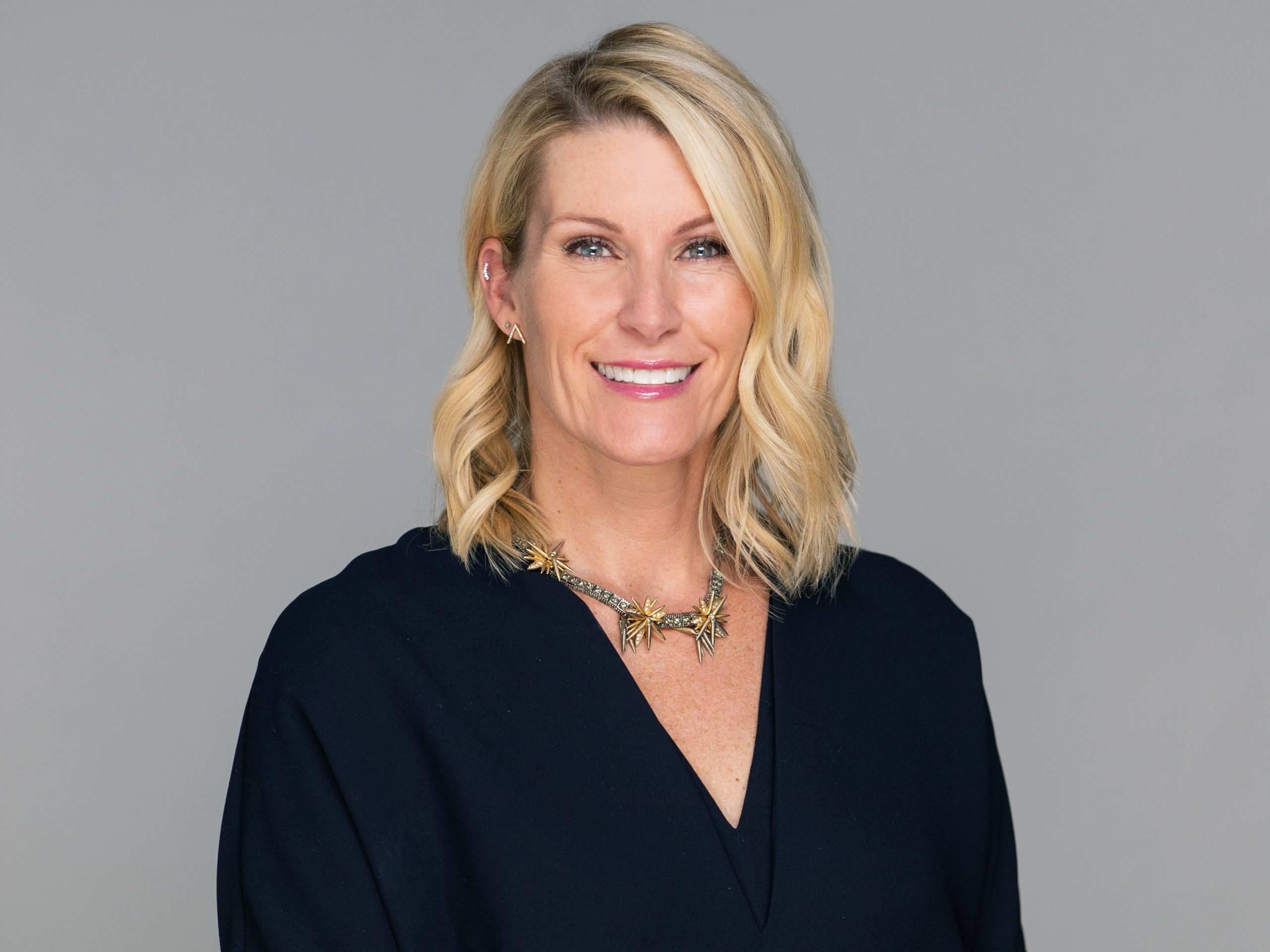 A headshot of Revlon CMO Martine Williamson wearing a black V-neck top, a silver and gold necklace, and stud earrings.