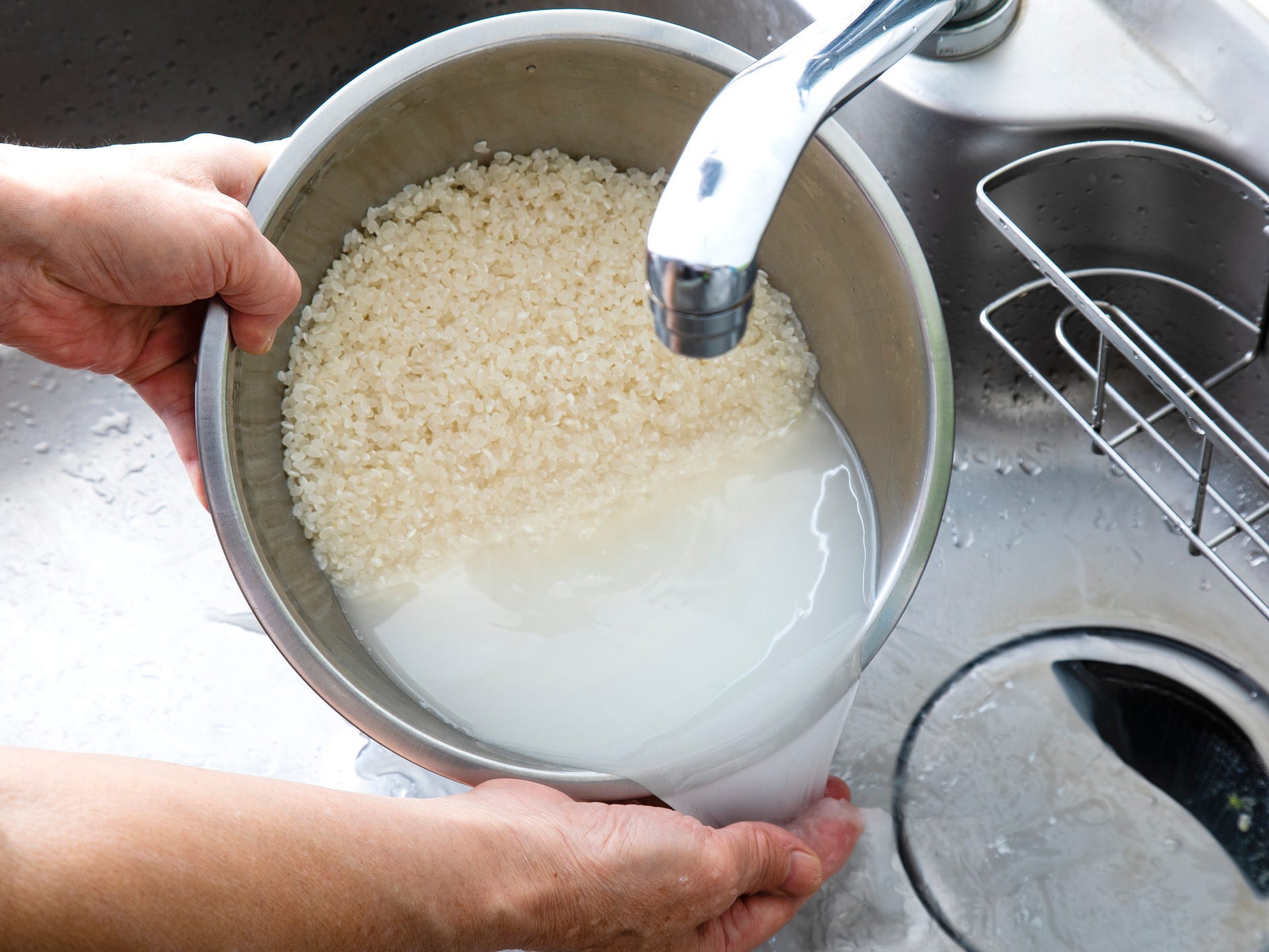 A person rinsing rice in a silver bowl under a kitchen faucet