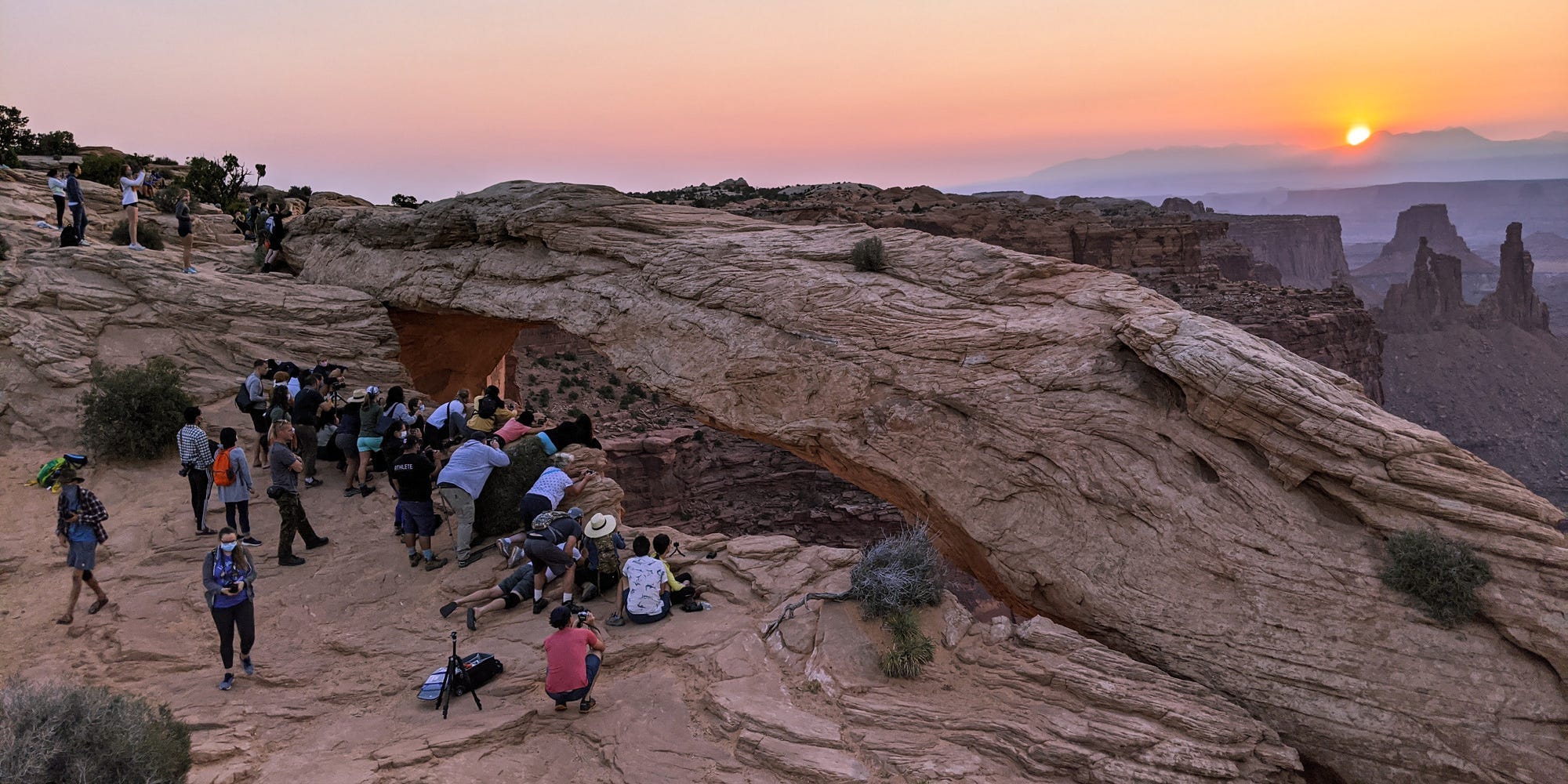 Mesa Arch, a red rock arch, at sunset with a crowd of people trying to take photos.