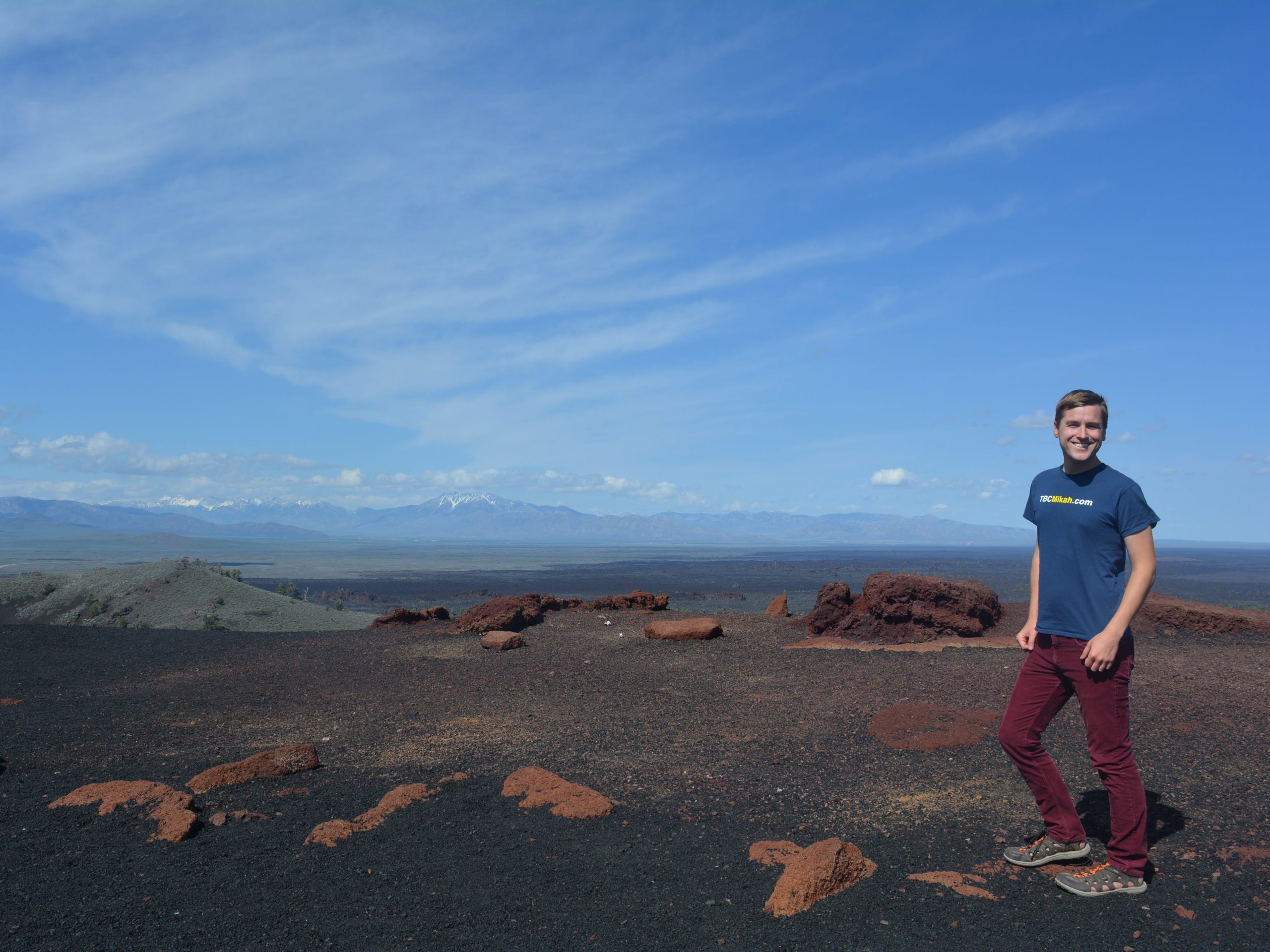 Mikah Meyer stands on the right side of a vast scene with black dirt, red rocks, and mountains in the background. The sky is blue with a few clouds.