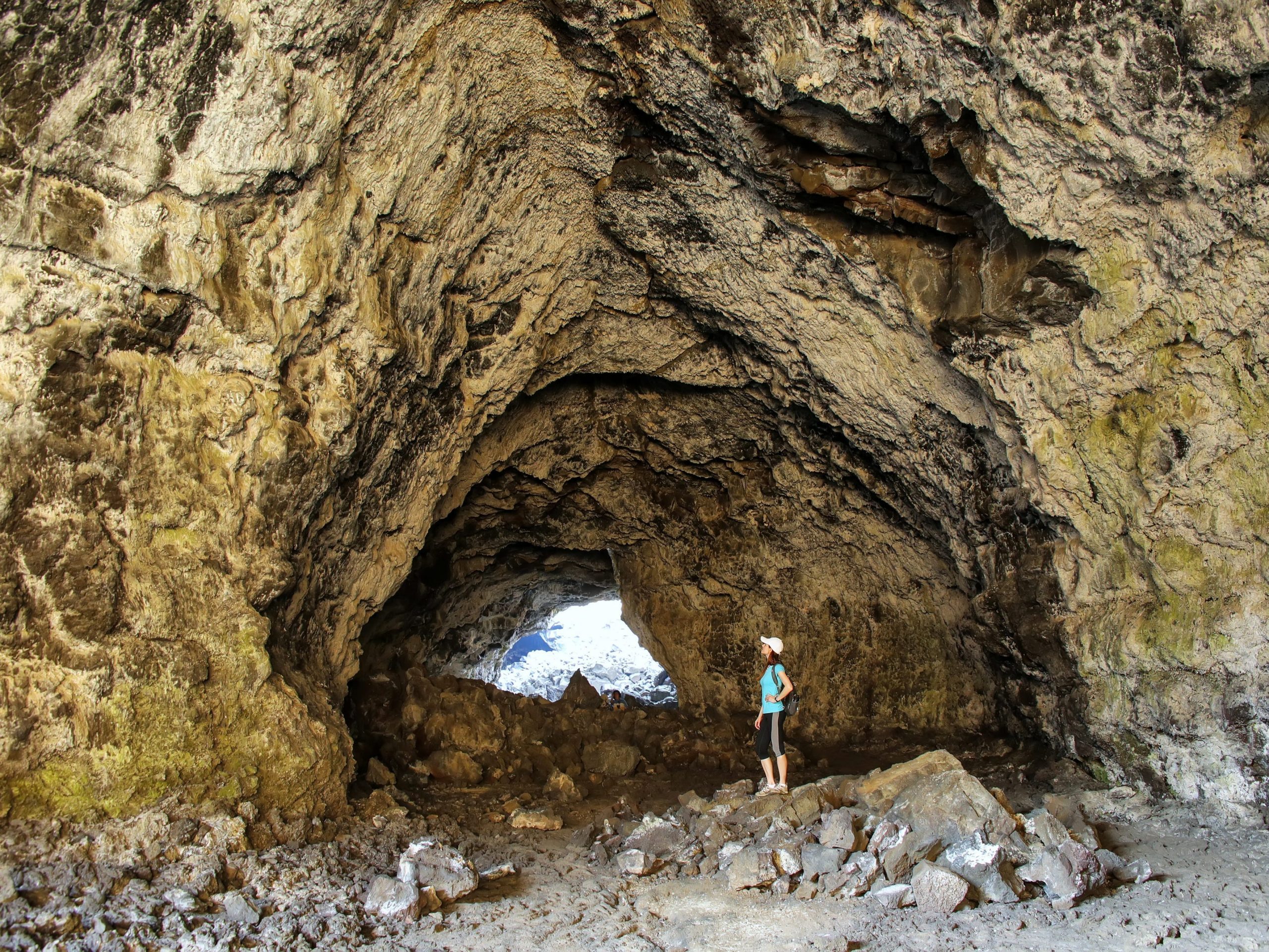A woman stands in a scene of a vast lava tunnel with light peering through from the other side.