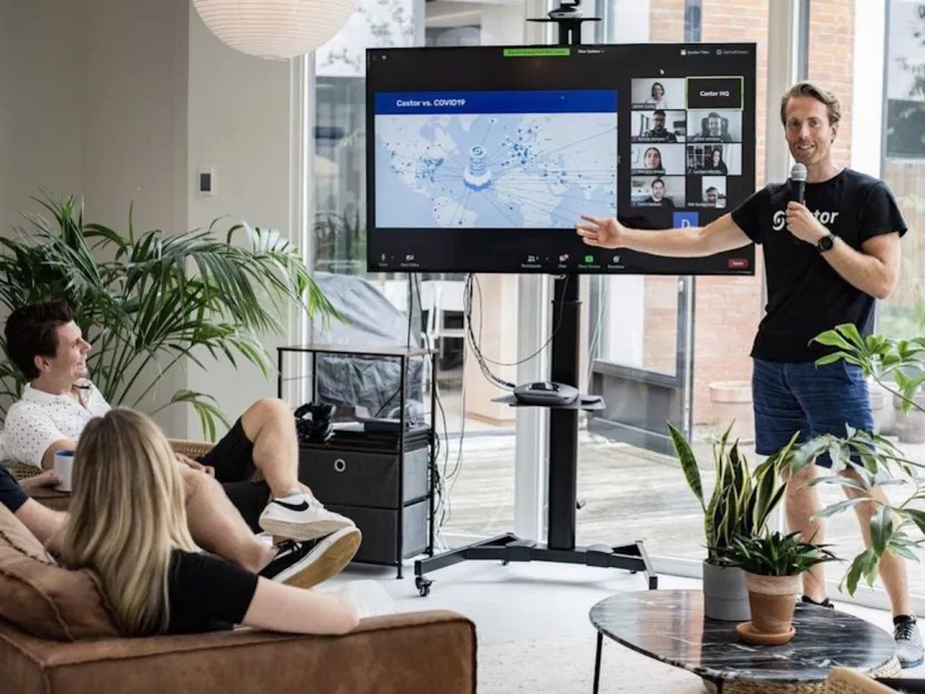 Castor CEO Derk Arts stands in front of a monitor with a slide presentation and teleconferencing participants as three people sitting on couches look on