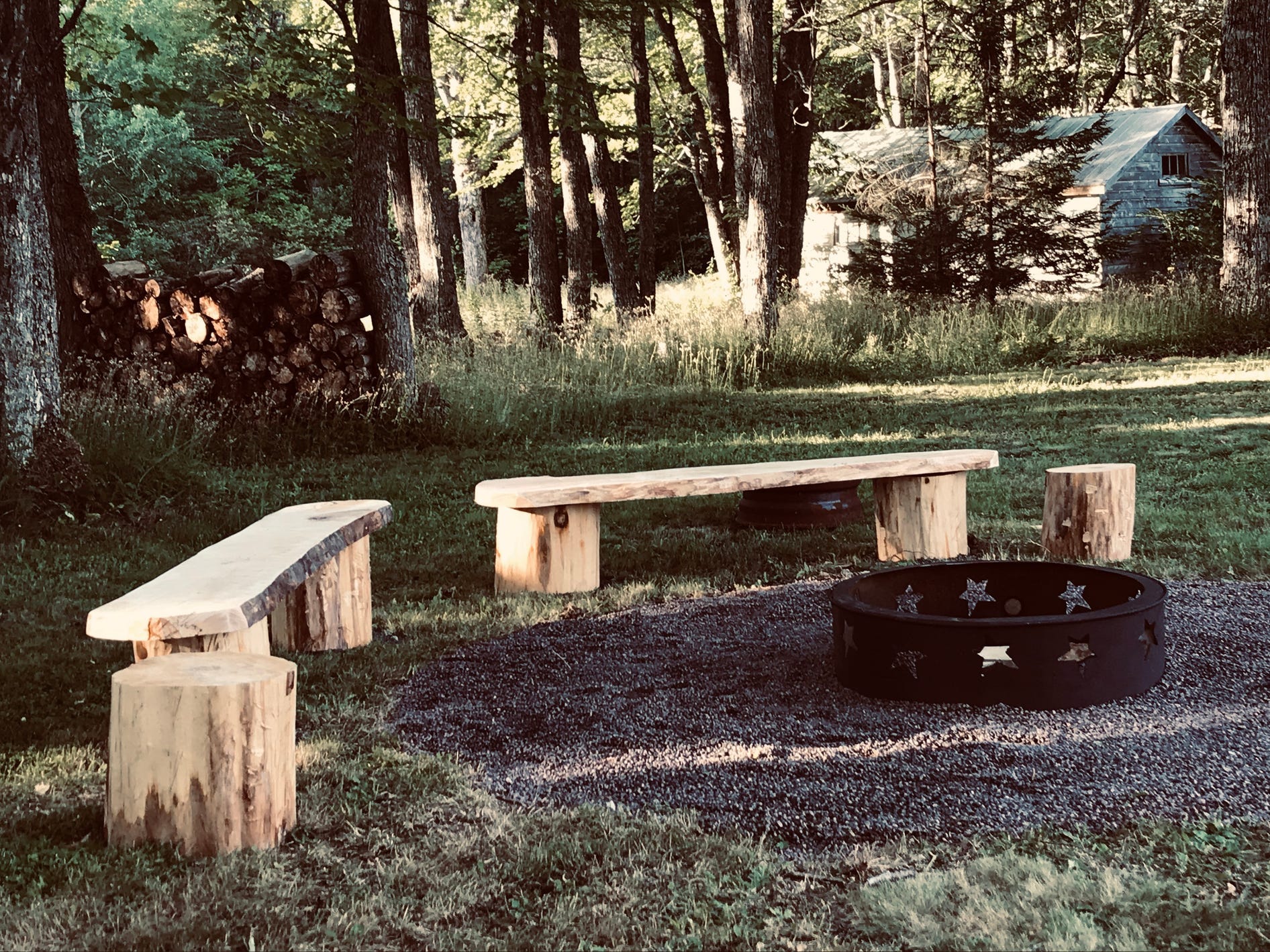 A fire pit in a wooded clearing surrounded by wooden benches and tree stump tables