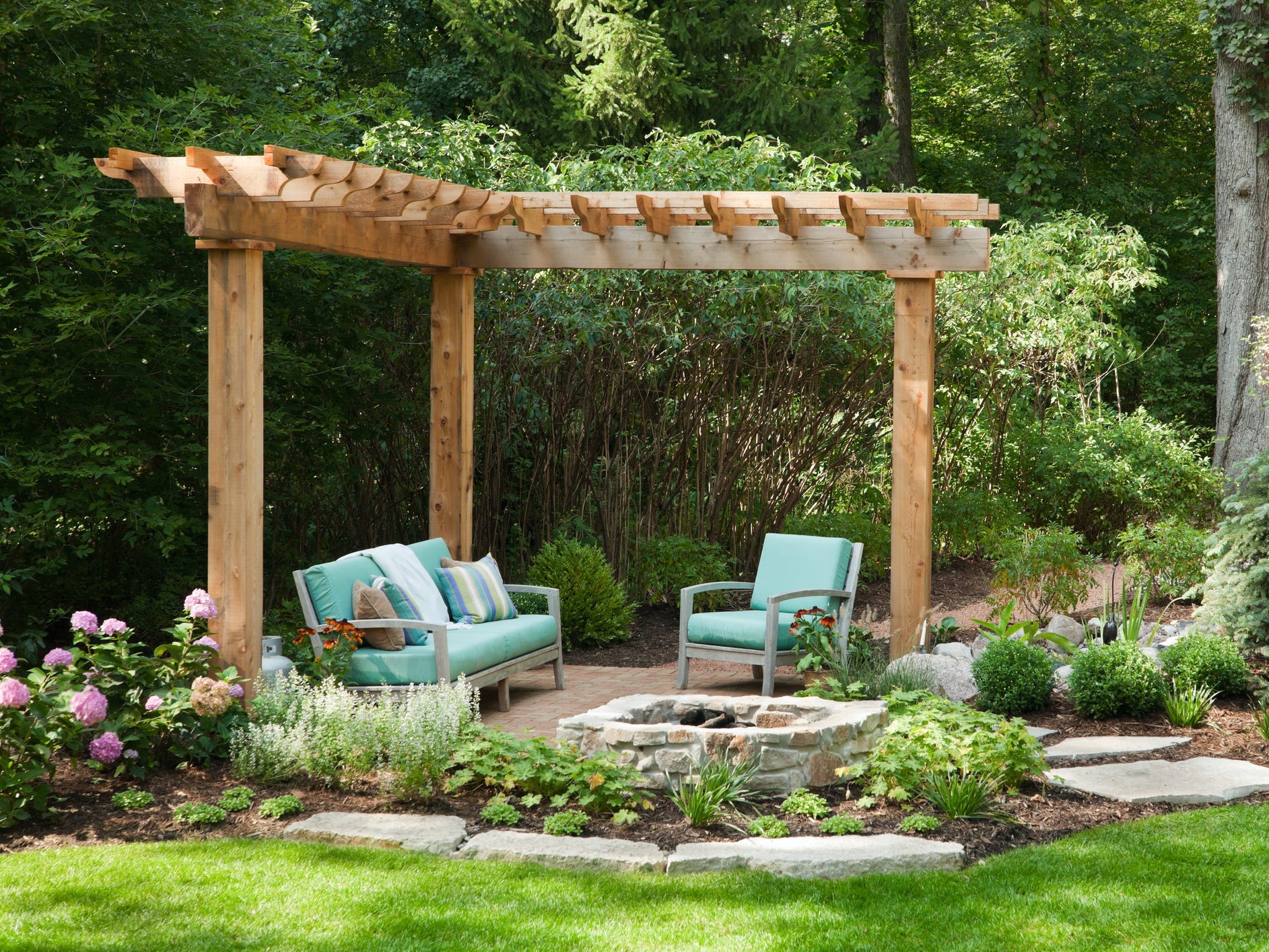A backyard with beautiful landscaping, a fire pit, blue cushioned couches, and a wooden pergola