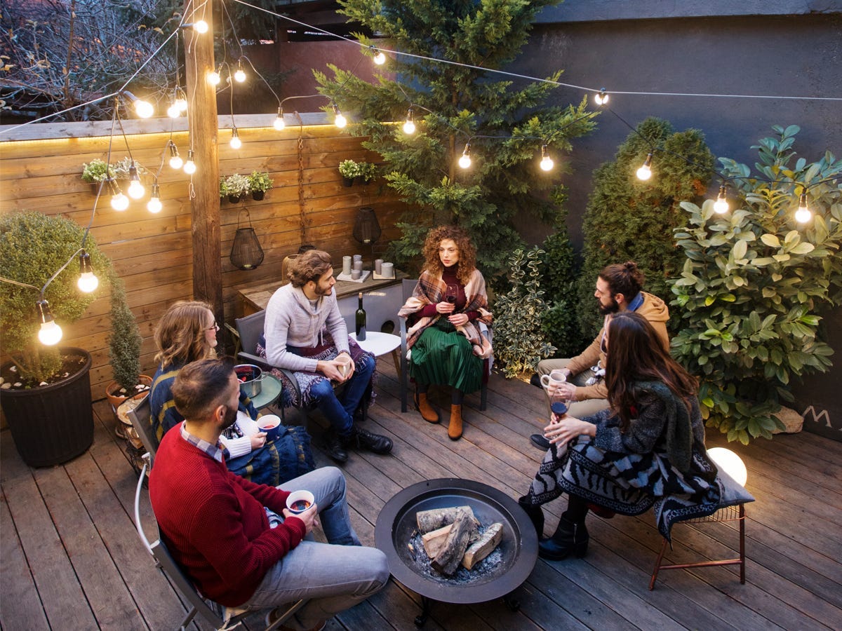 A group of friends sitting around a backyard fire pit with string lights overhead