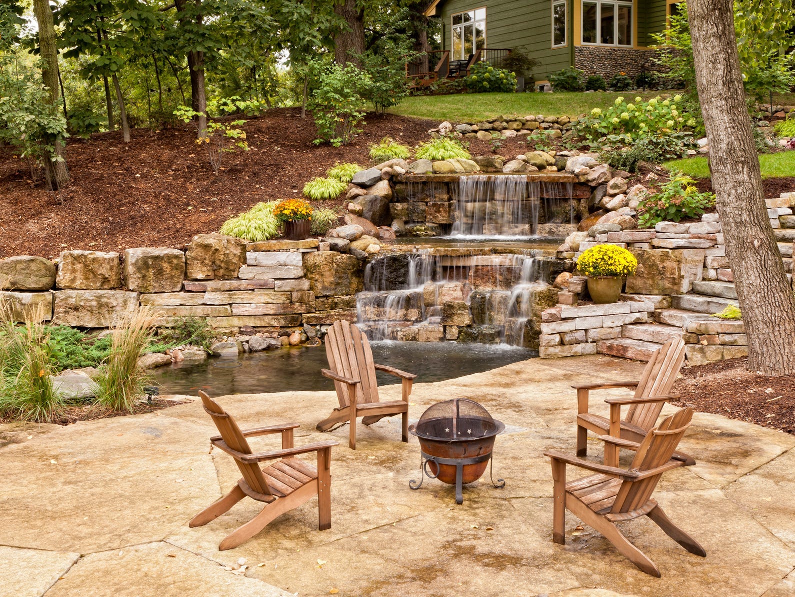 A large backyard with a stonework waterfall and a small portable fire pit surrounded by Adirondack chairs